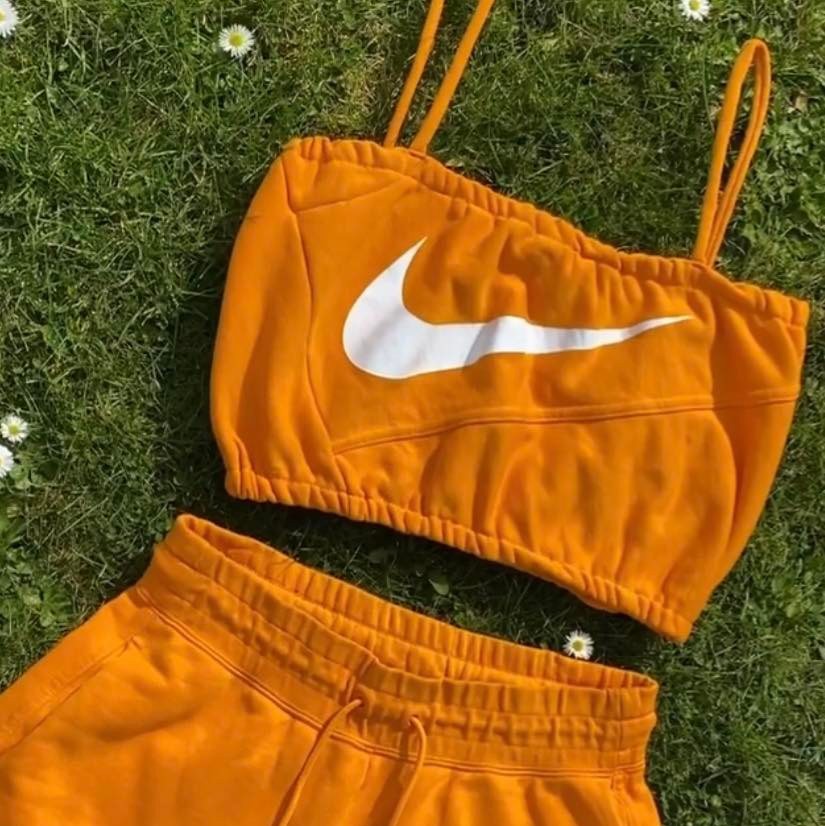 bright orange crop top with straps and big white nike swoosh logo on the the front with matching shorts on a background of grass with small white daisies