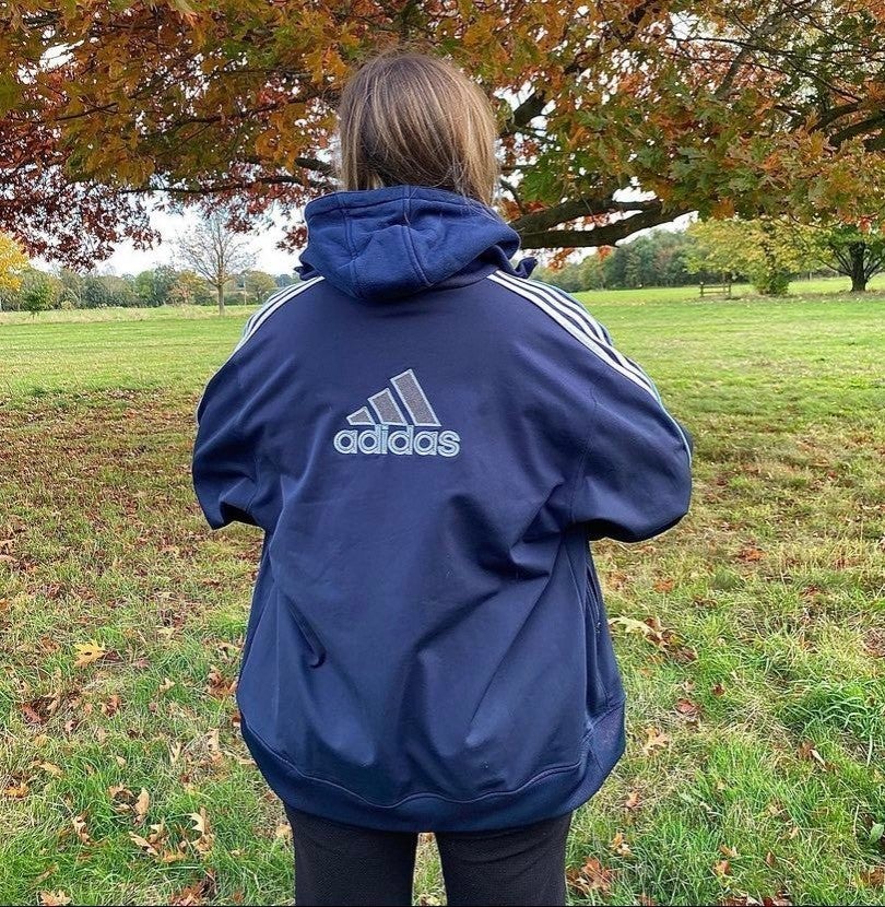 the back of a navy blue adidas jacket with big text adidas logo and symbol worn on a model with brown hair and background of grass and trees