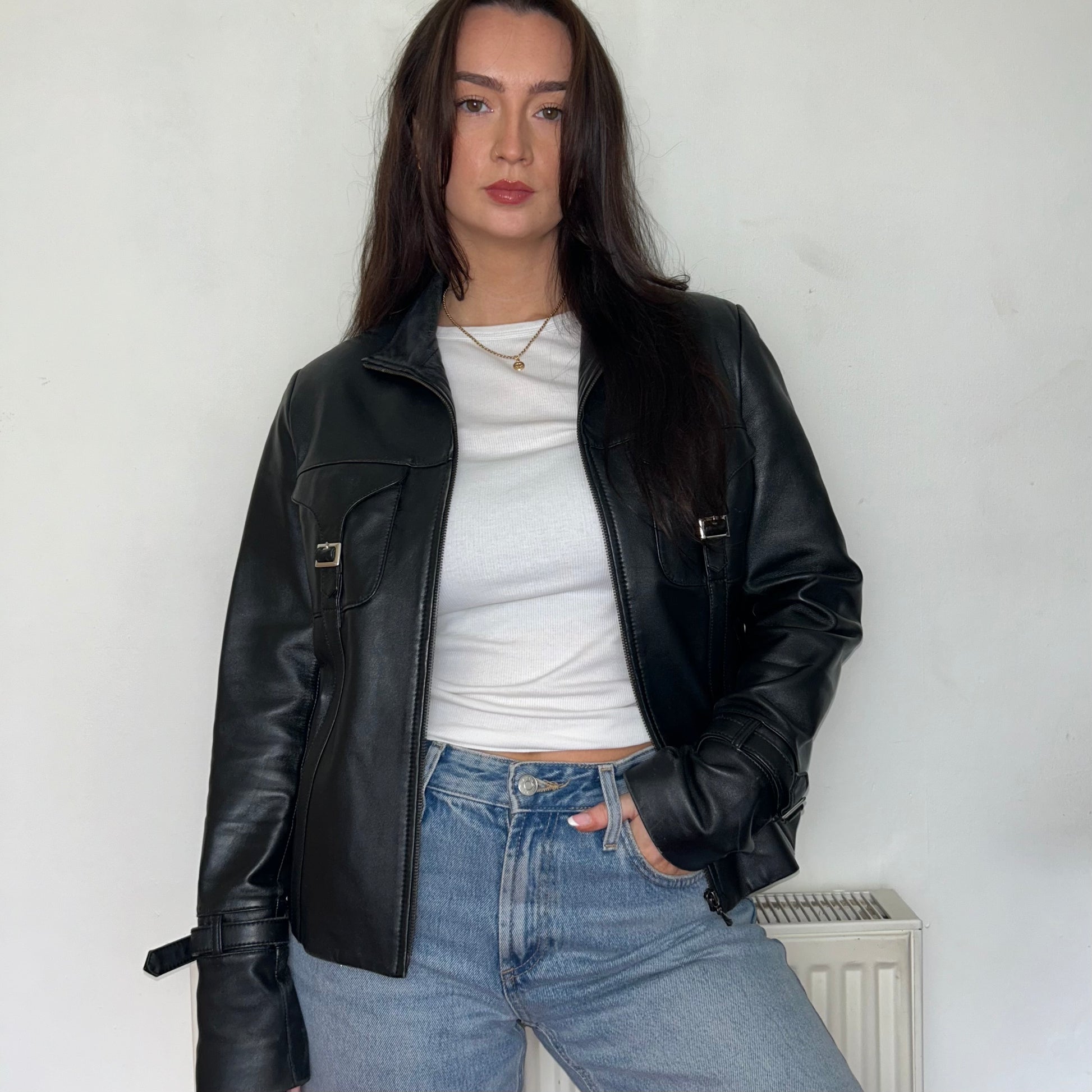 black leather buckle bomber jacket shown on a model wearing a white crop top and blue jeans with hand in pocket
