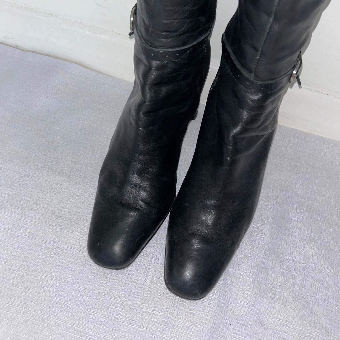 toes of black knee high leather buckle boots on a white background