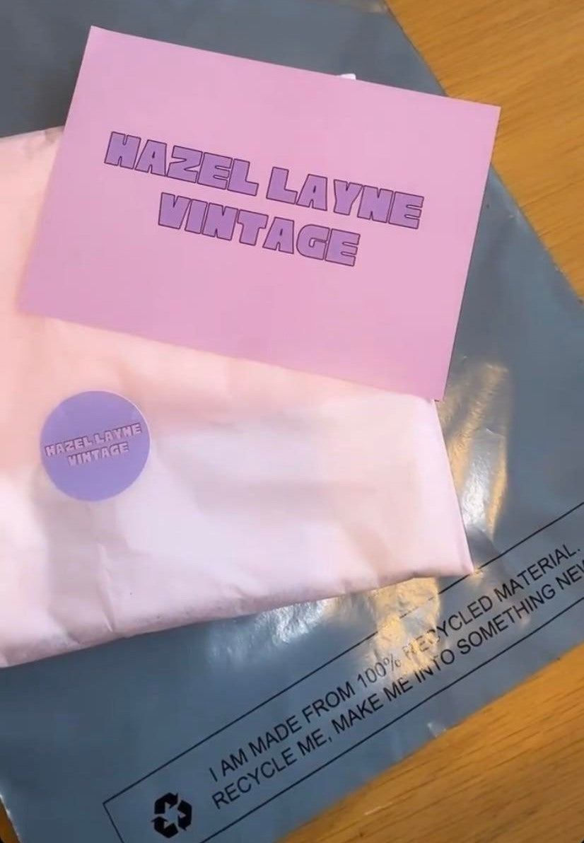 a light pink postcard with hazel layne vintage text writing on it on top of an item wrapped in tissue paper with a hazel layne vintage sticker on top of a grey mailers bag on a brown table