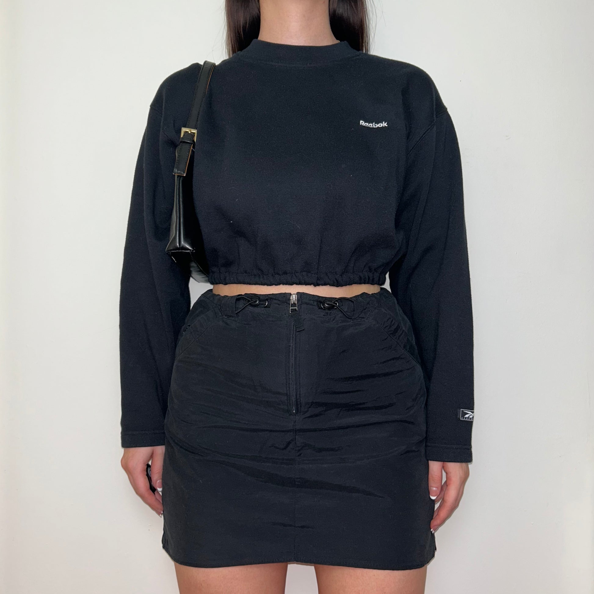 black cropped sweatshirt with white reebok logo shown on a model wearing a black mini skirt and black shoulder bag