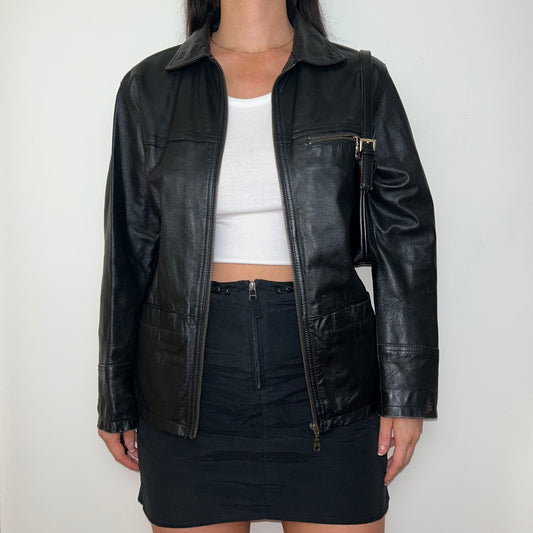 black real leather bomber jacket shown on a model wearing a white crop top and black mini skirt with a black shoulder bag