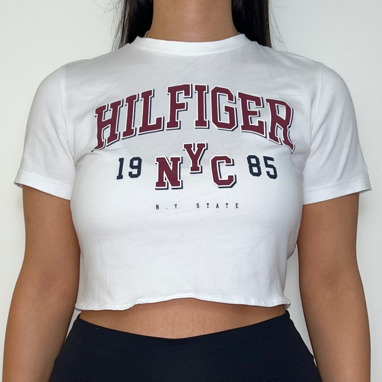 close up of white short sleeve crop top with hilfiger logo shown on a model wearing black trousers