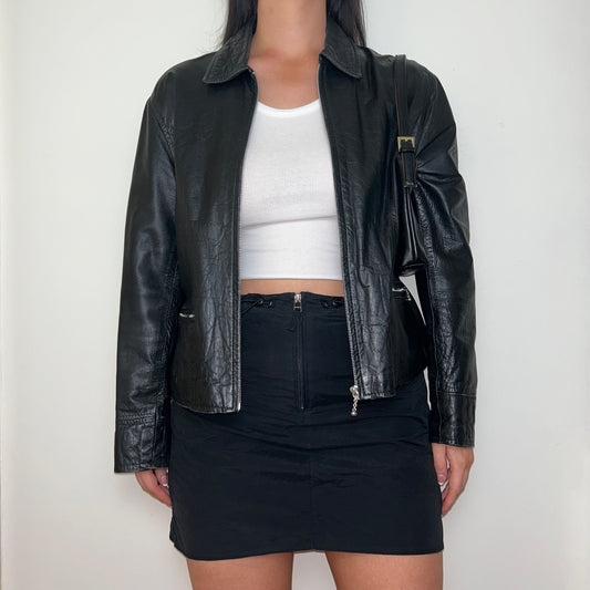 black leather zip up jacket shown on a model wearing a white crop top and black mini skirt with a black shoulder bag
