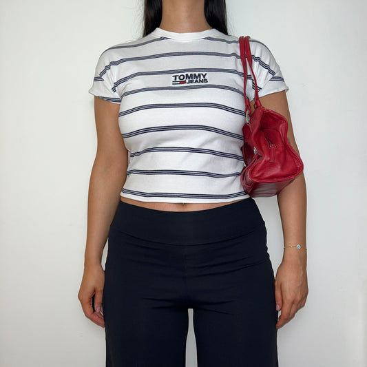 white stripe short sleeve crop top with black tommy jeans logo shown on a model wearing black trousers and a red shoulder bag