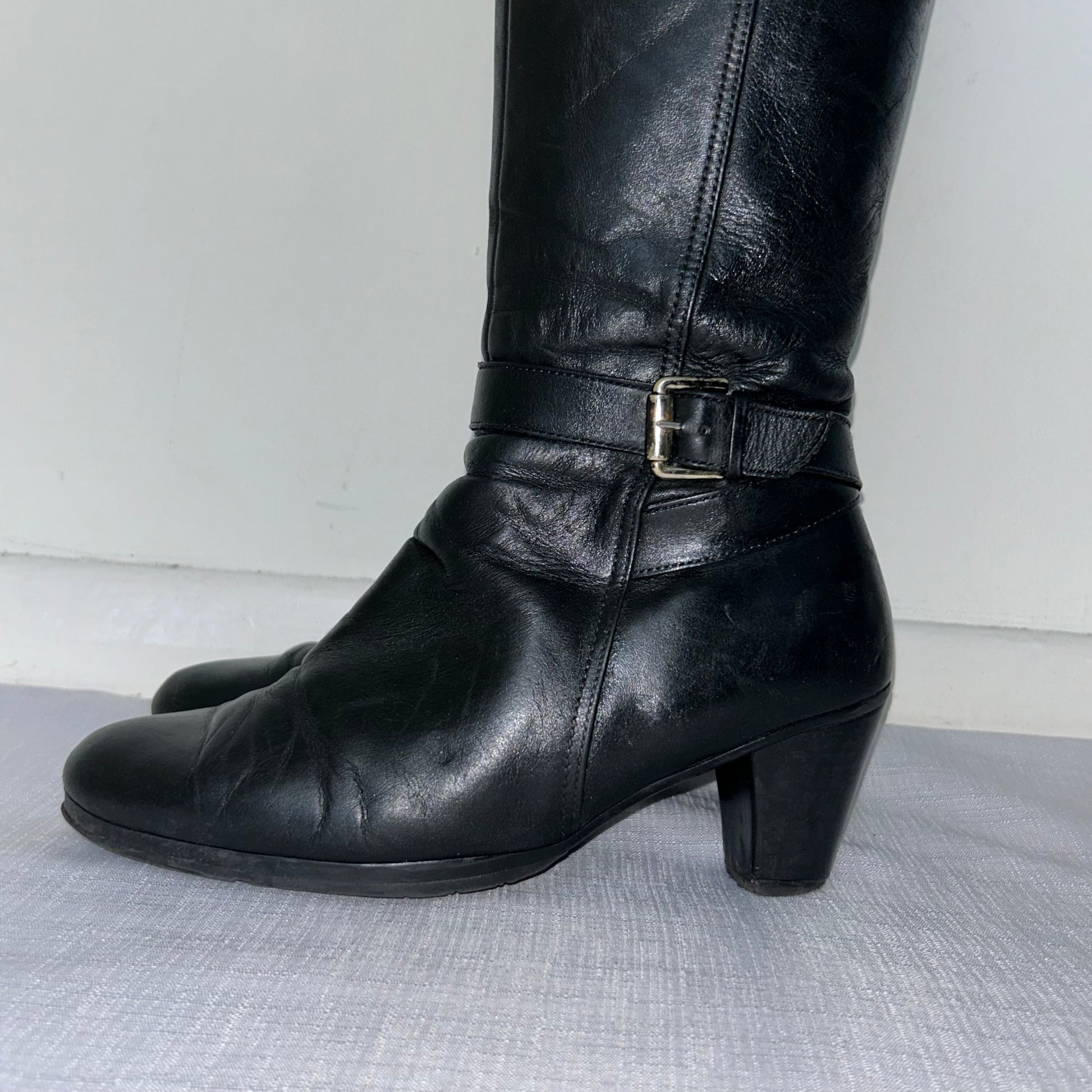 close up of black knee high silver buckle boots shown on a white background
