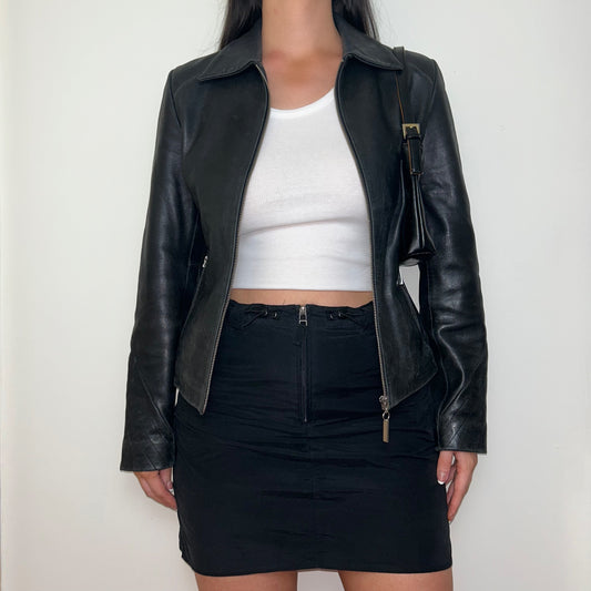 black real leather jacket shown on a model wearing a white crop top and black mini skirt with a black shoulder bag