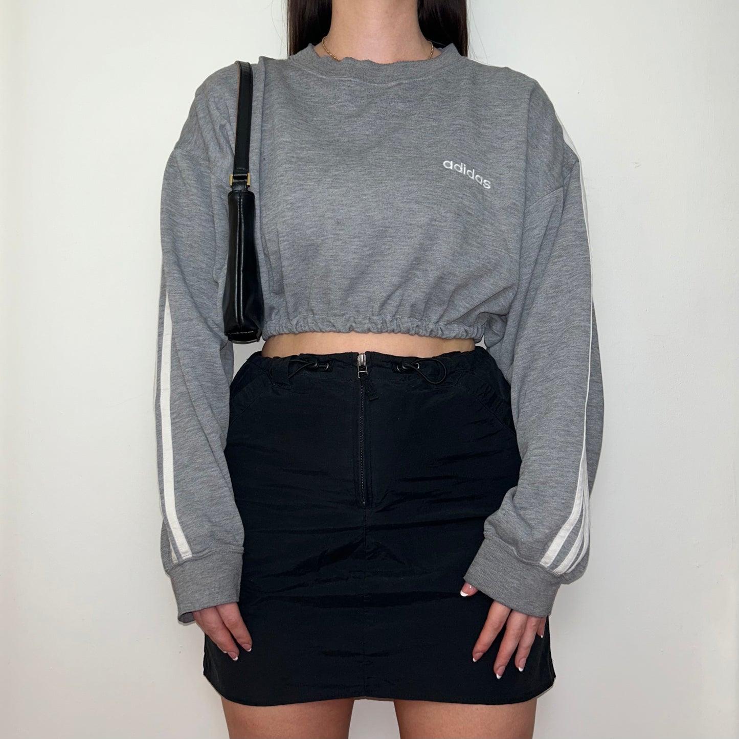 grey cropped sweatshirt with white adidas logo shown on a model wearing a black mini skirt and black shoulder bag