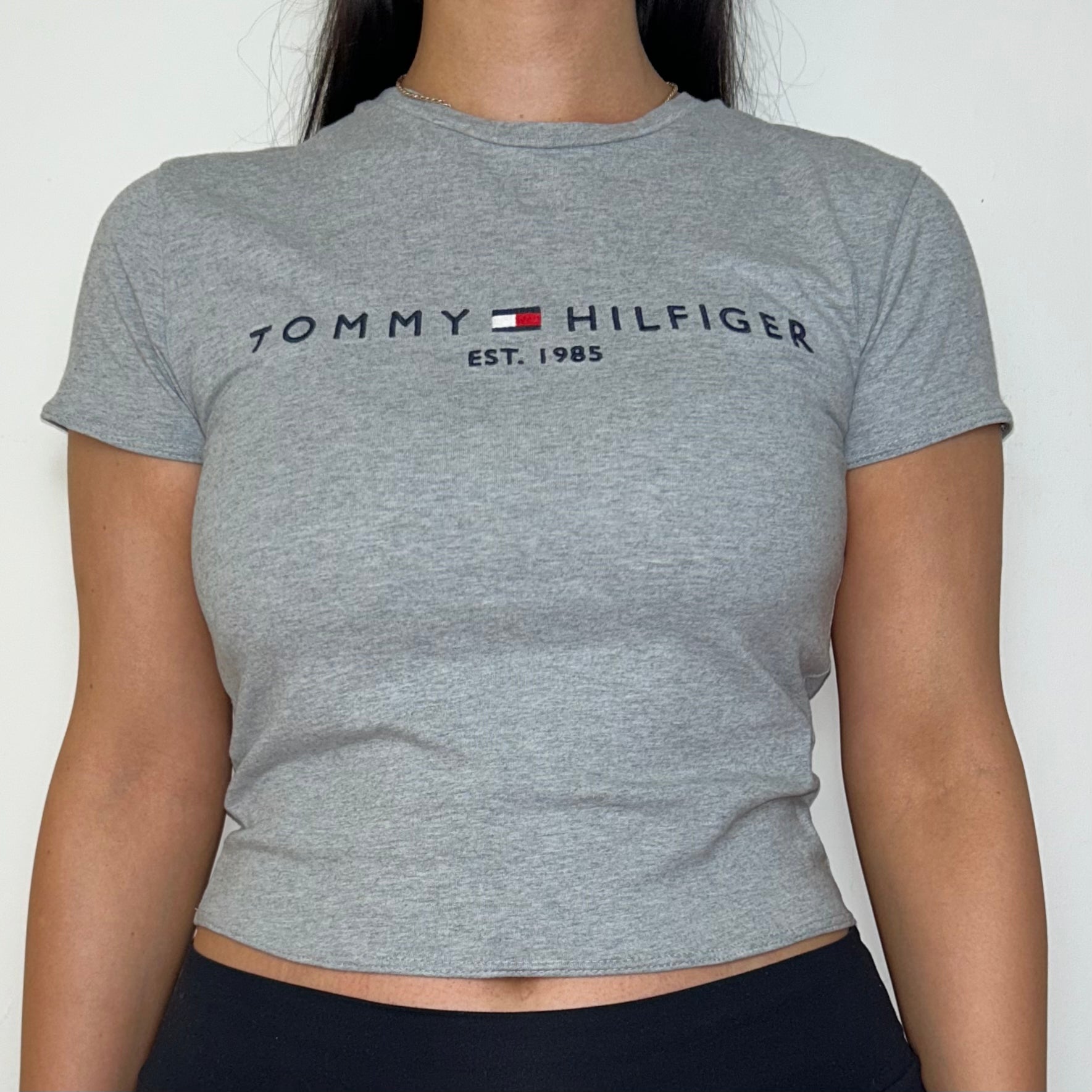 close up of grey short sleeve crop top with black tommy hilfiger logo shown on a model wearing black trousers