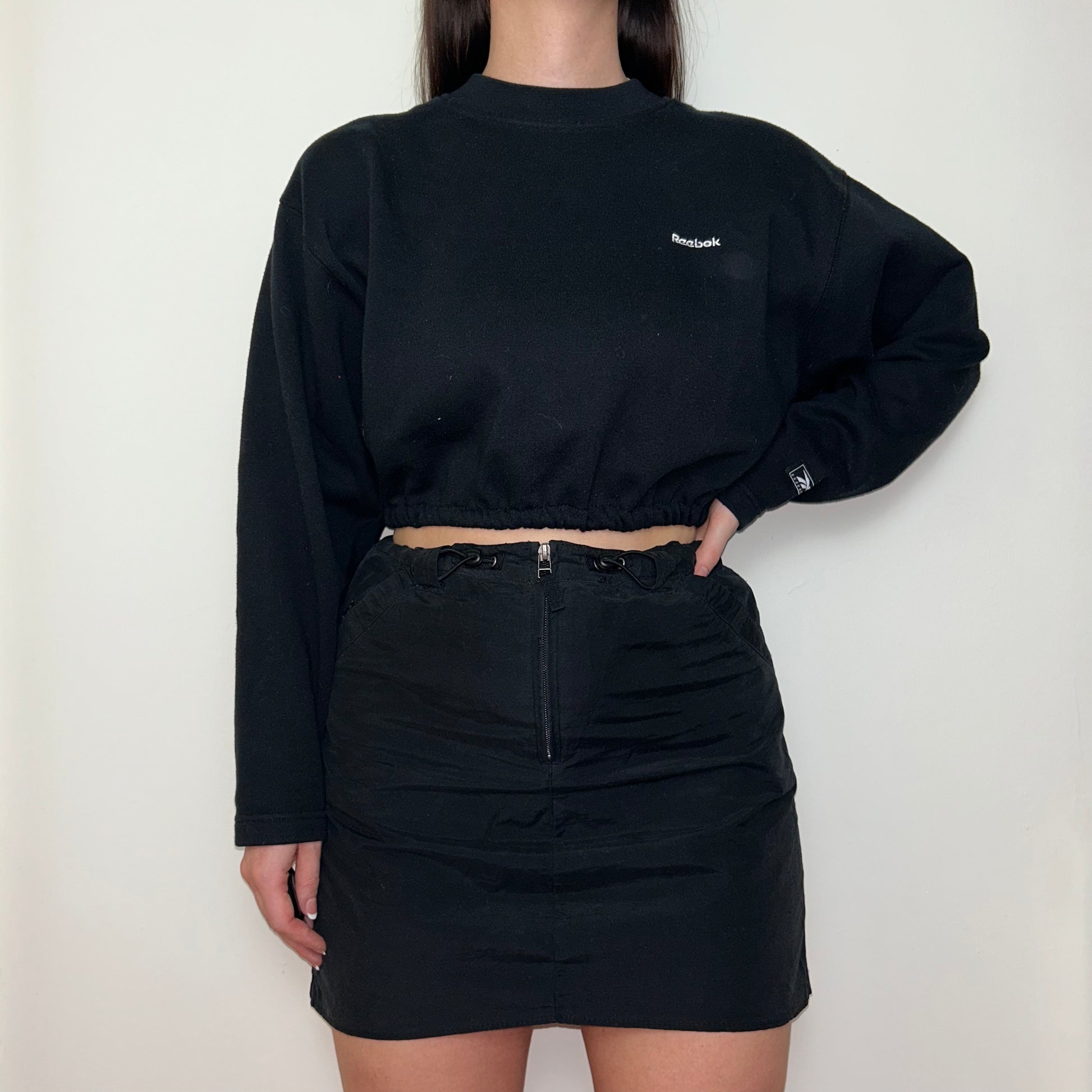 black cropped sweatshirt with white reebok logo shown on a model wearing a black mini skirt