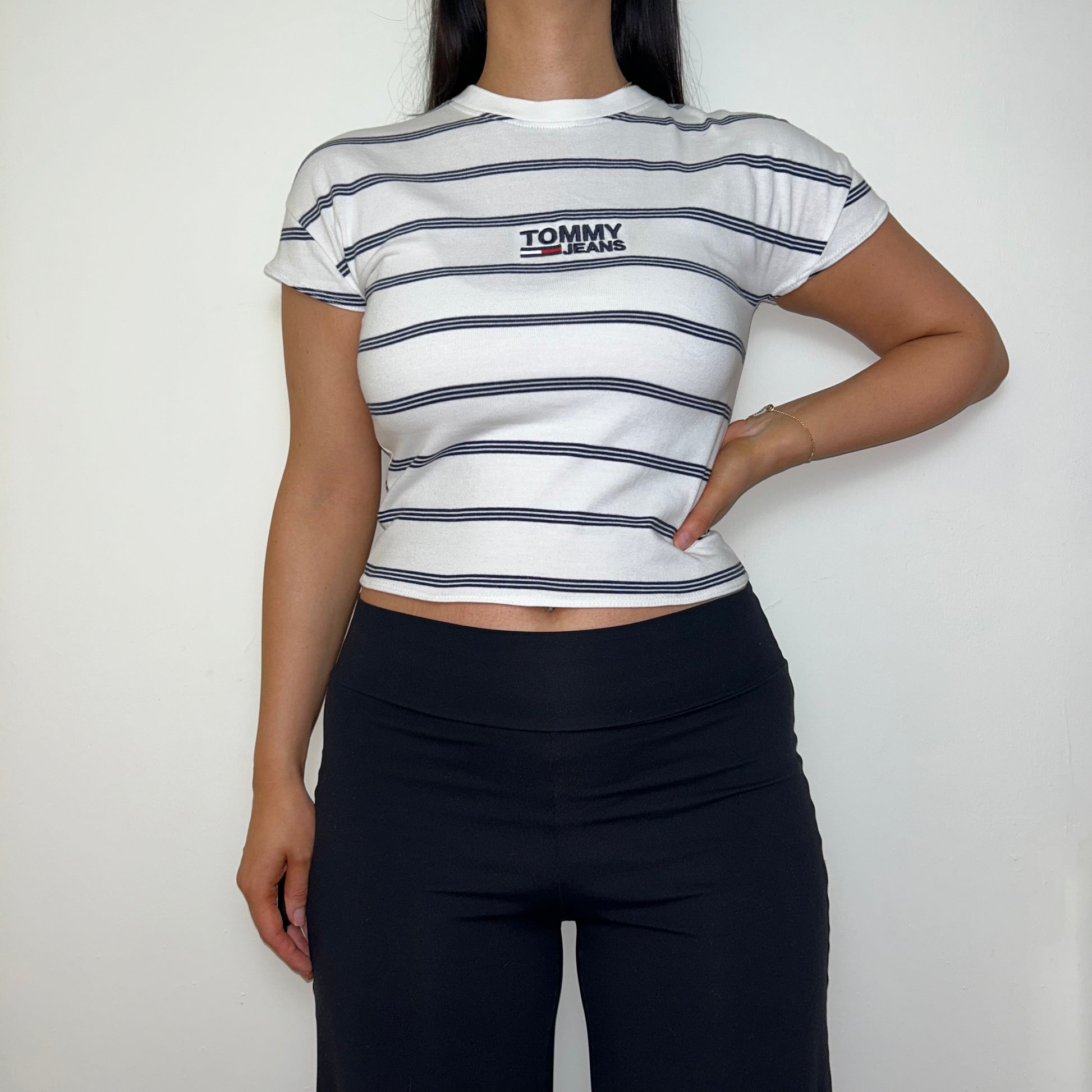 white stripe short sleeve crop top with black tommy jeans logo shown on a model wearing black trousers