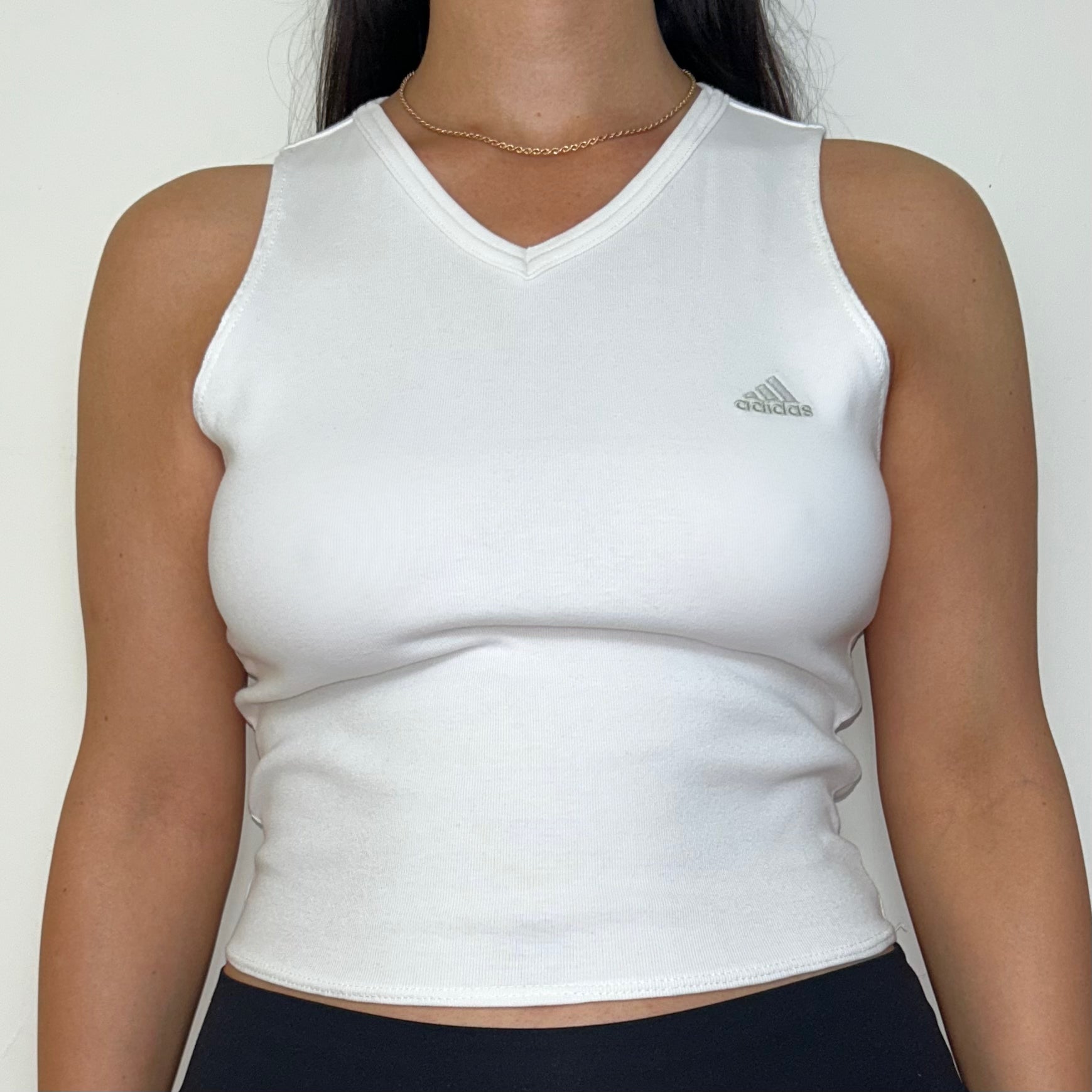 close up of white sleeveless crop top with grey adidas logo shown on a model wearing black trousers