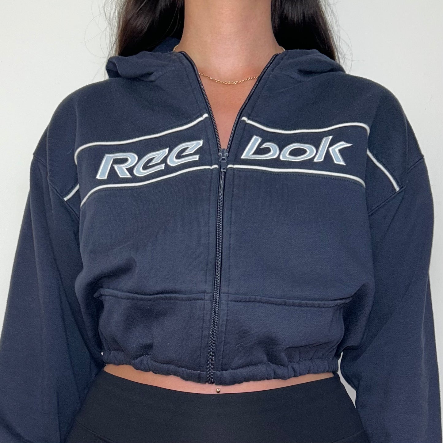 close up of navy cropped hoodie with white reebok logo shown on a model wearing black trousers