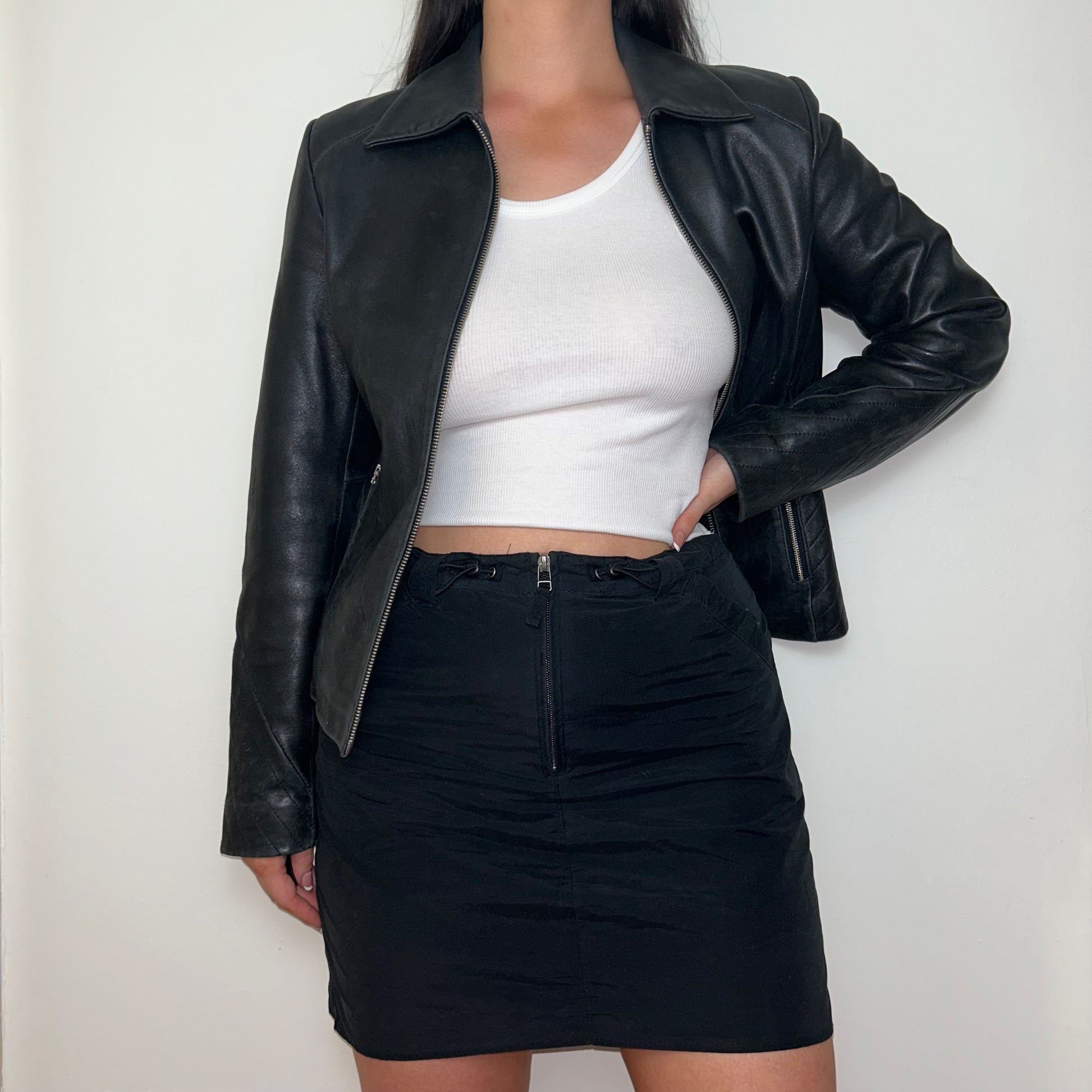 black real leather jacket shown on a model wearing a white crop top and black mini skirt