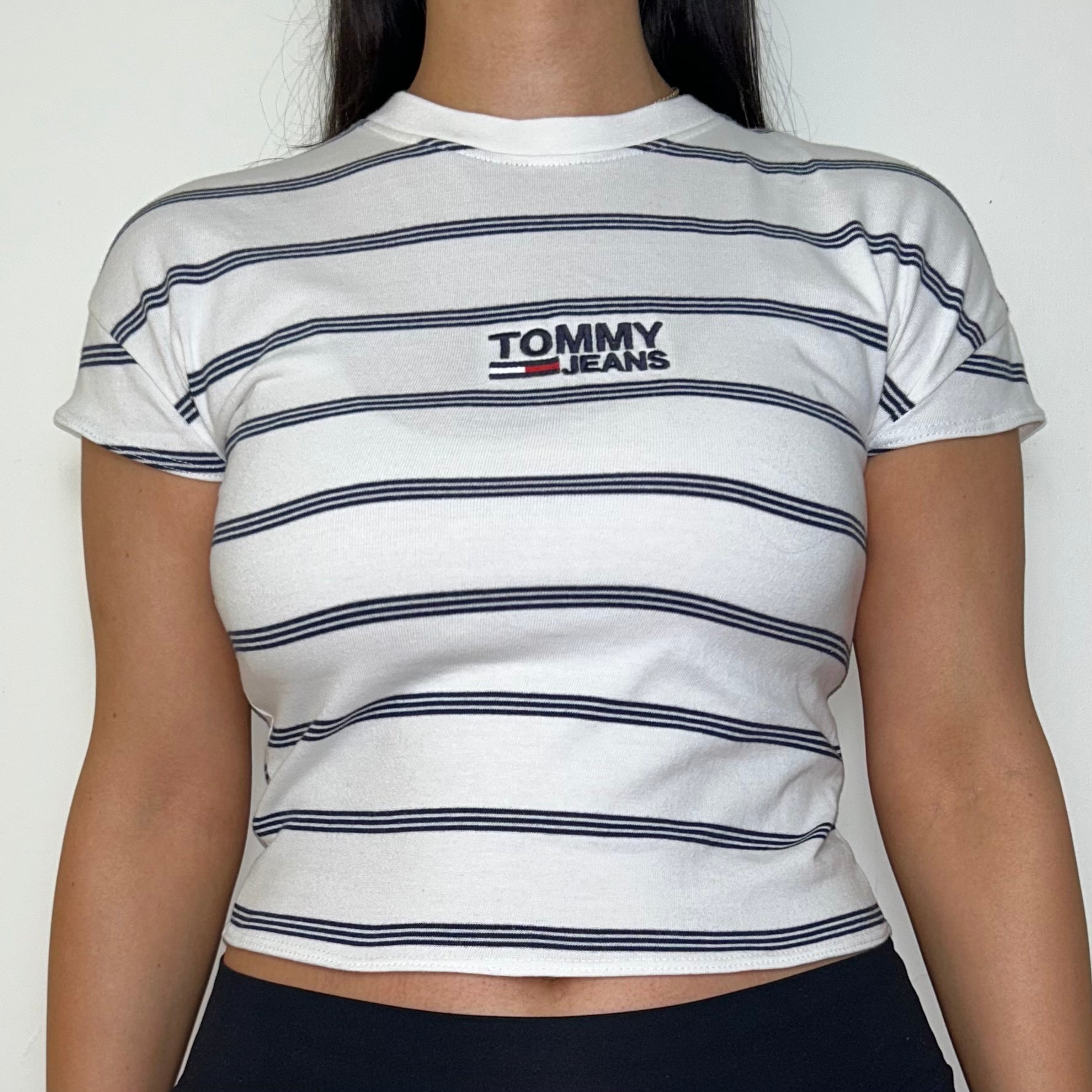 close up of white stripe short sleeve crop top with black tommy jeans logo shown on a model wearing black trousers