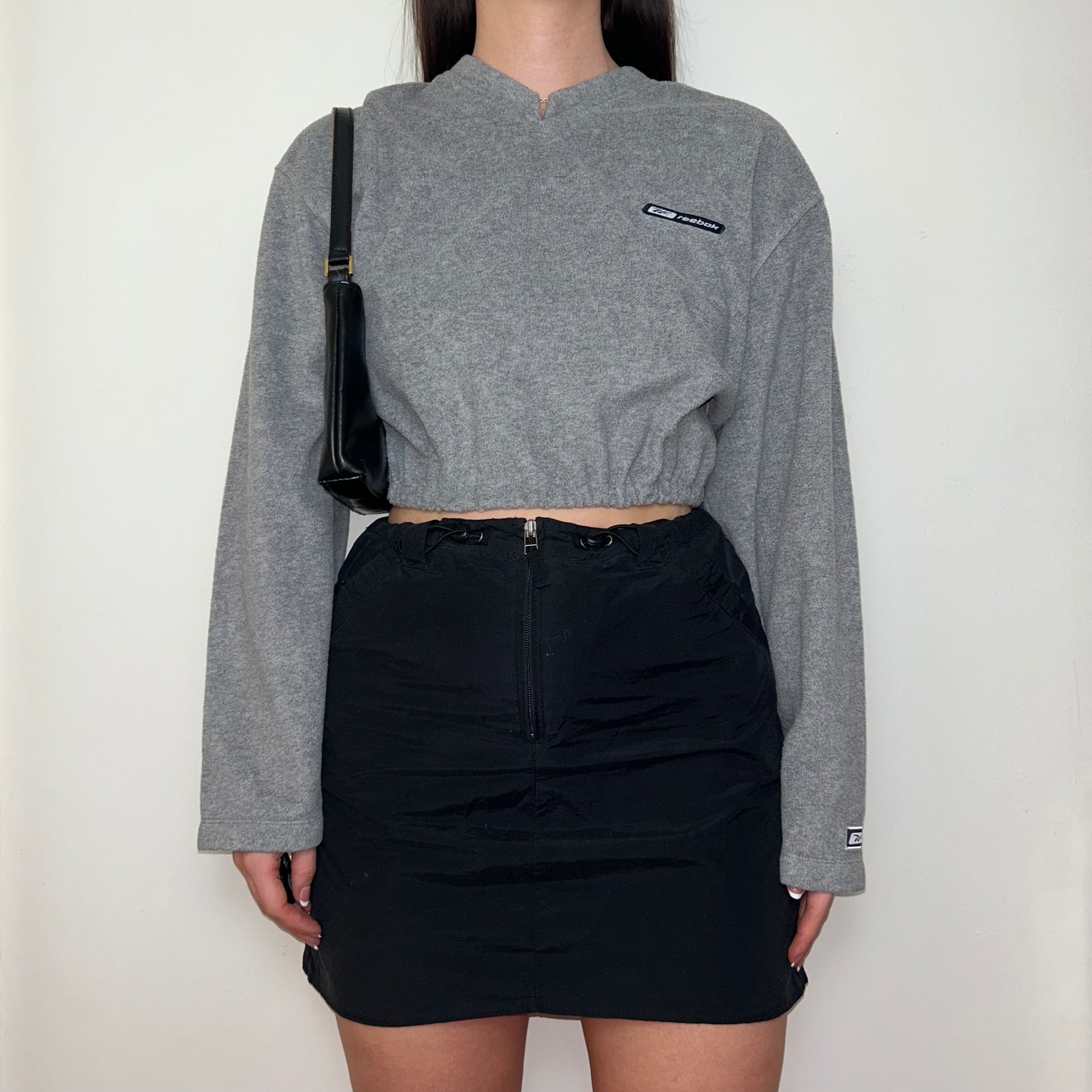 grey cropped sweatshirt with reebok logo shown on a model wearing a black skirt and black shoulder bag
