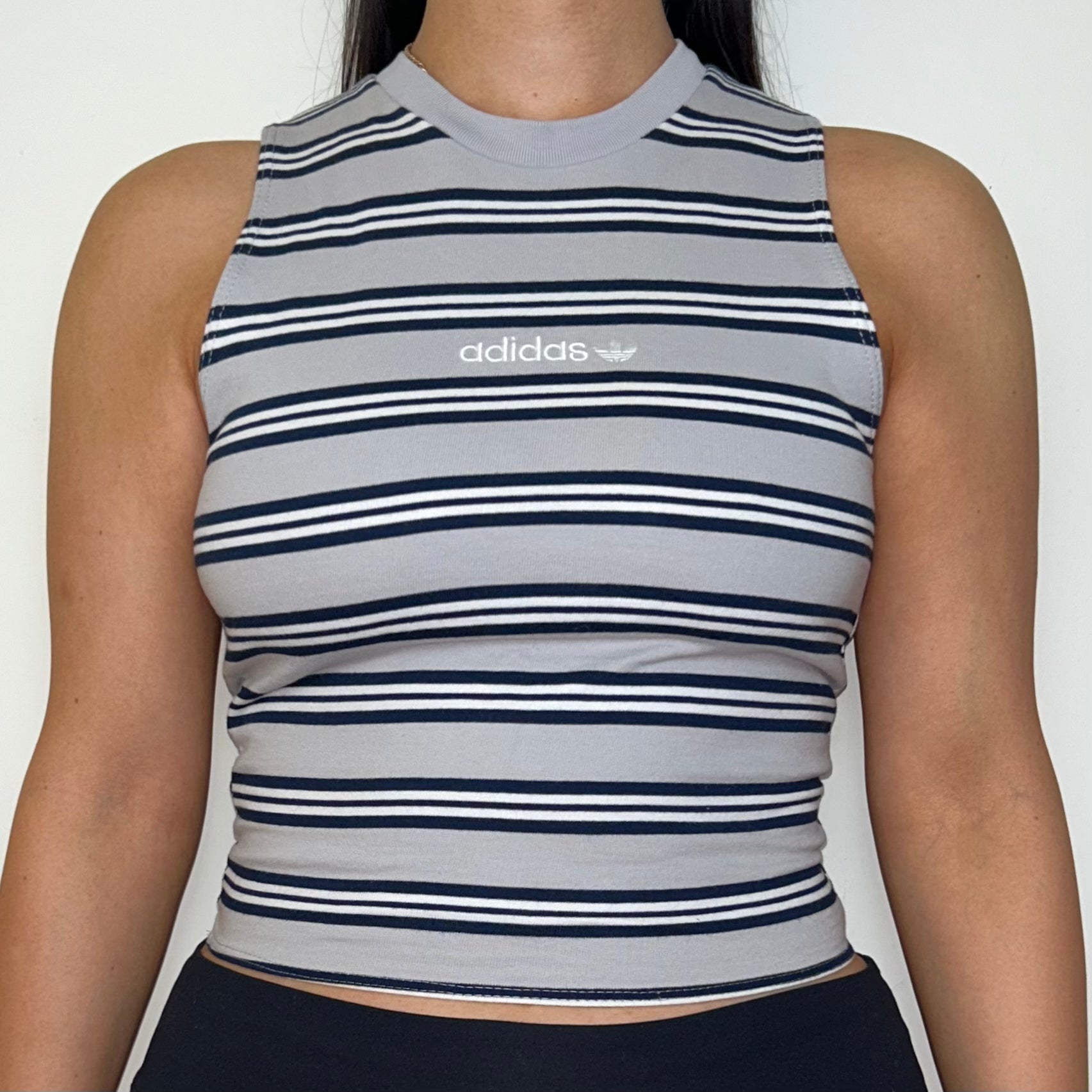 close up of grey stripe sleeveless crop top with white adidas logo shown on a model wearing black trousers