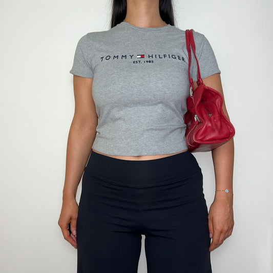 grey short sleeve crop top with black tommy hilfiger logo shown on a model wearing black trousers and a red shoulder bag