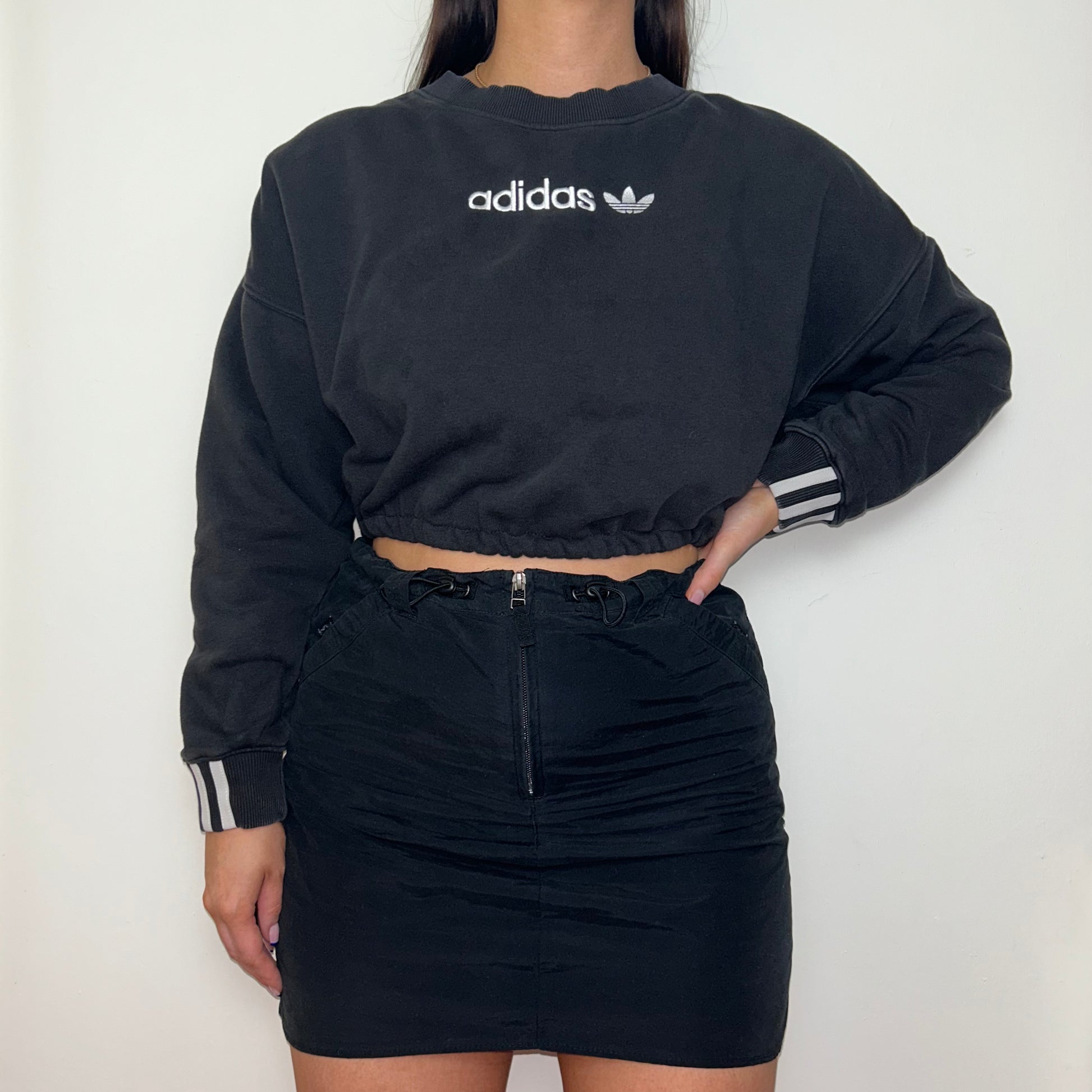 black cropped sweatshirt with white adidas logo shown on a model wearing a black mini skirt