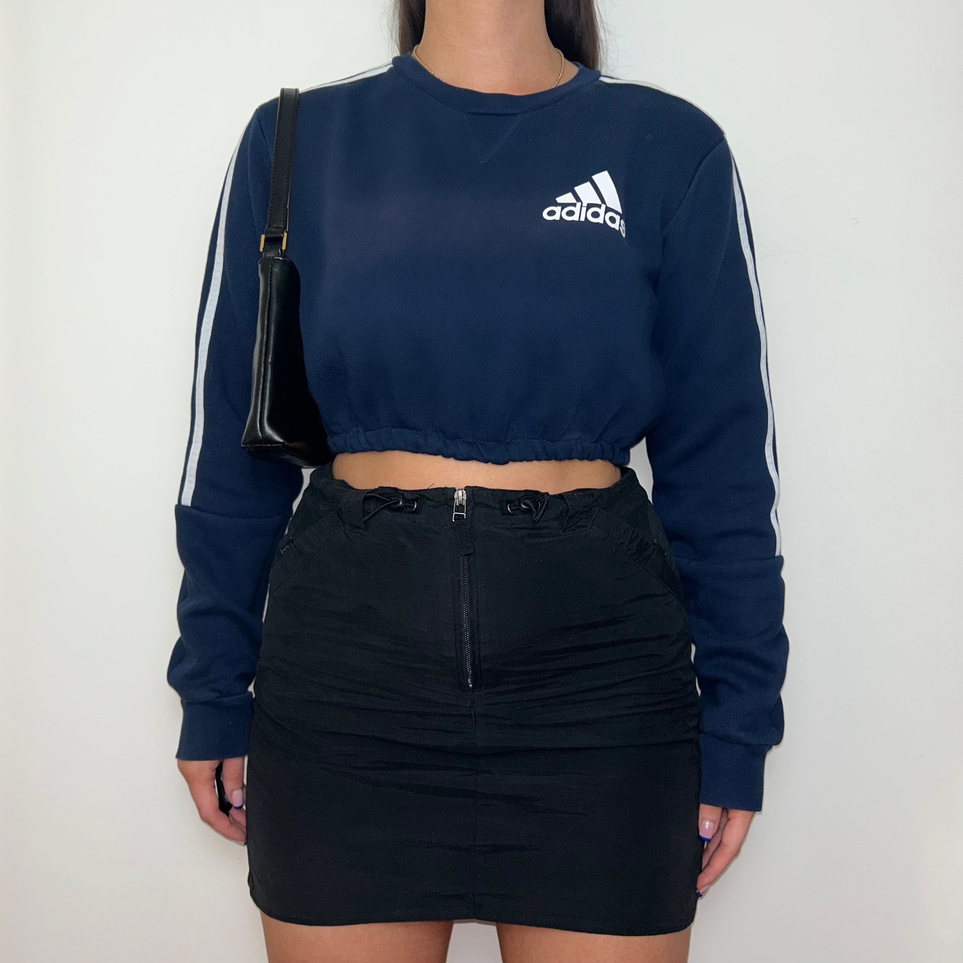navy cropped sweatshirt with white adidas logo shown on a model wearing a black mini skirt and black shoulder bag