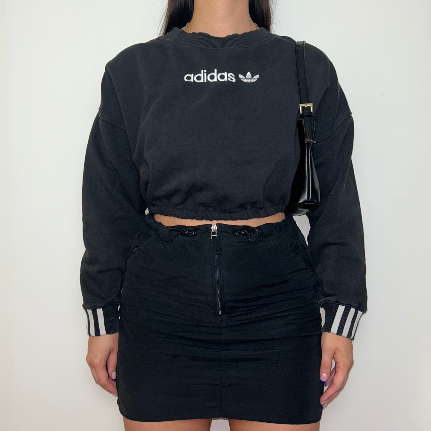black cropped sweatshirt with white adidas logo shown on a model wearing a black mini skirt and black shoulder bag