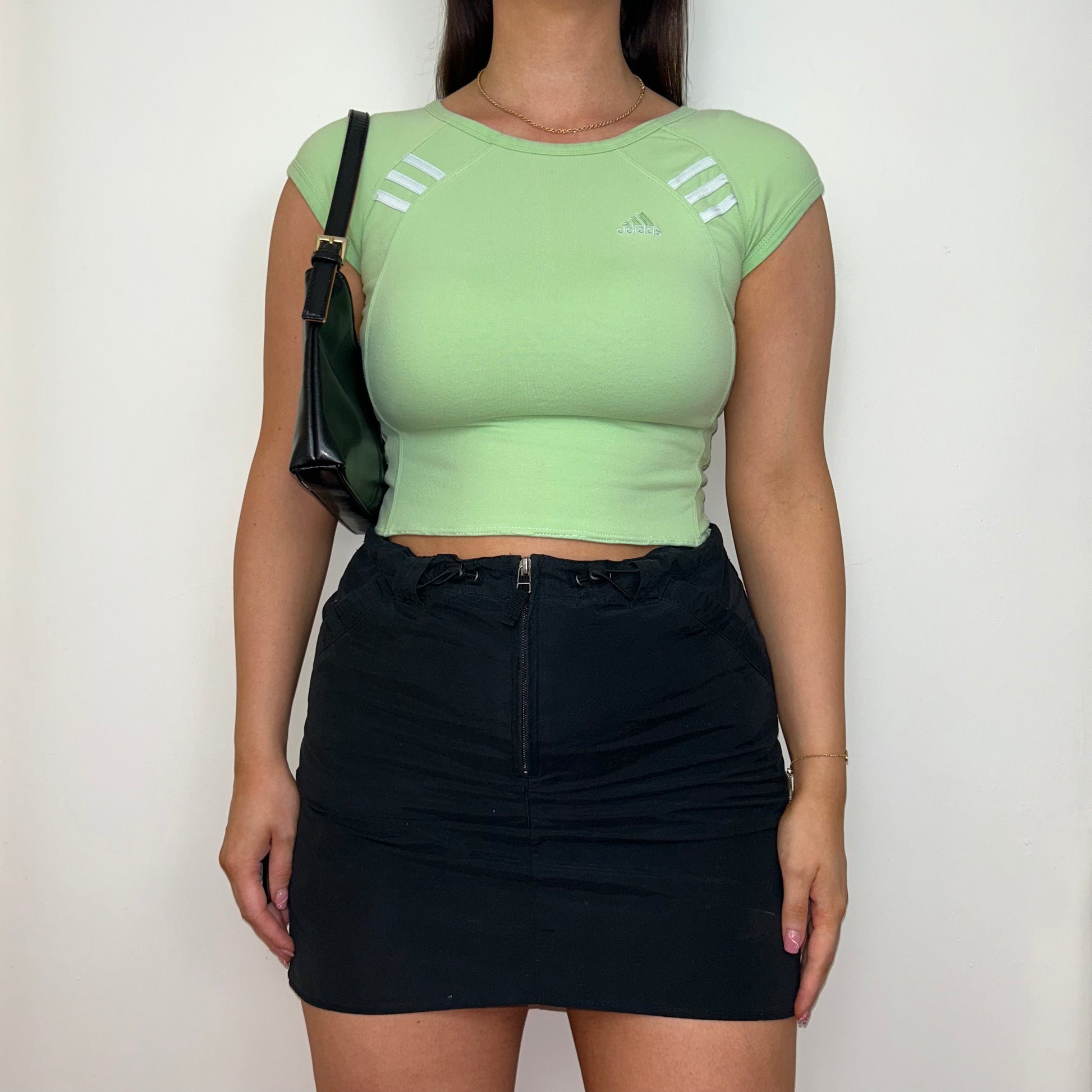 light green short sleeve crop top with white adidas logo shown on a model wearing a black mini skirt with a black shoulder bag