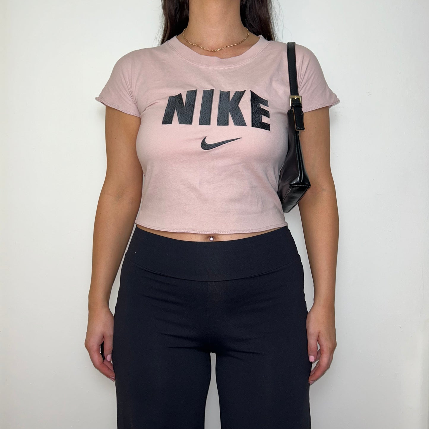 light pink short sleeve crop top with black nike logo shown on a model wearing black trousers and a black shoulder bag