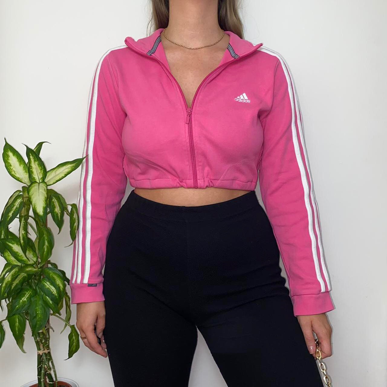 bright pink cropped jumper with small white Adidas text and symbol logo on chest shown on a model wearing black trousers