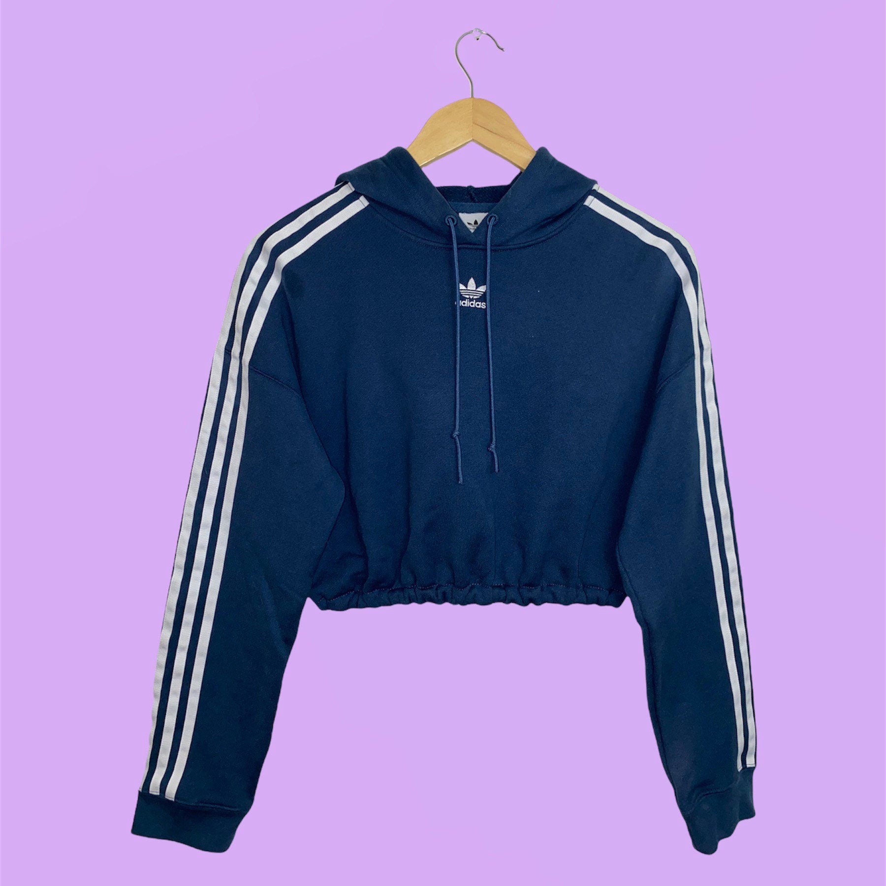 navy cropped hoodie with small Adidas text logo and symbol and white 3 stripes down the sleeves shown on a wood clothes hanger on a lilac background