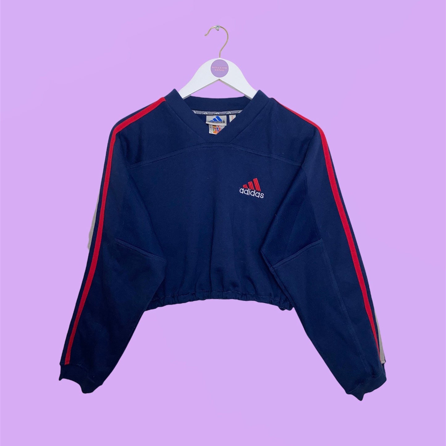 navy v neck cropped sweatshirt with red small adidas text and white symbol logo shown on a white clothes hanger on a lilac background