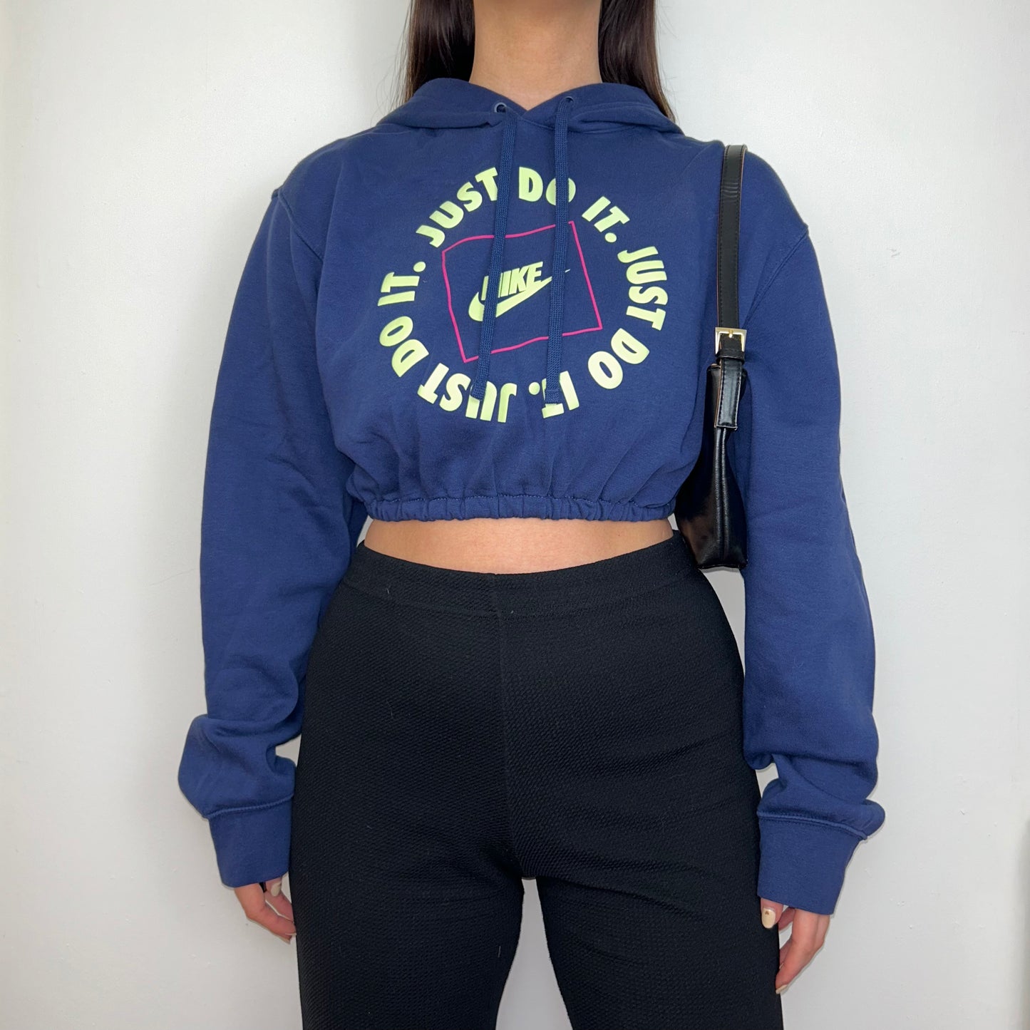 navy blue cropped hoodie with yellow and pink nike text logos shown on a model wearing black trousers and a black shoulder bag