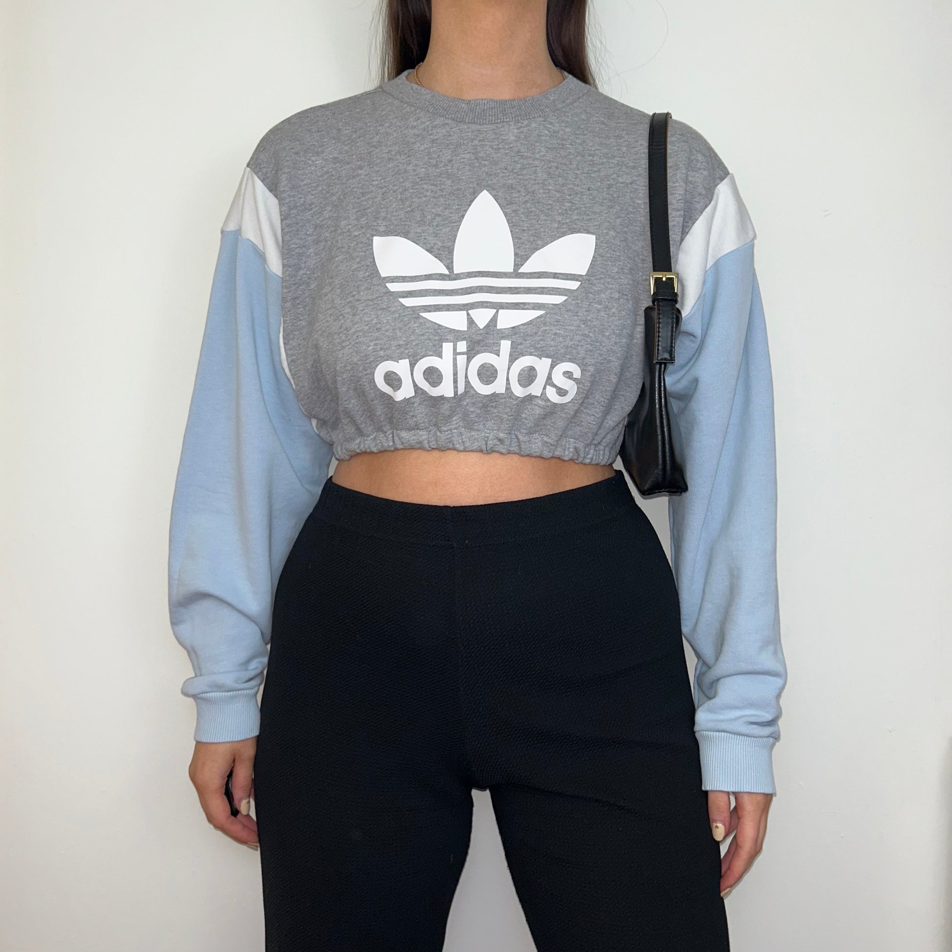 grey and blue cropped sweatshirt with white adidas logo shown on a model wearing black trousers and a black shoulder bag