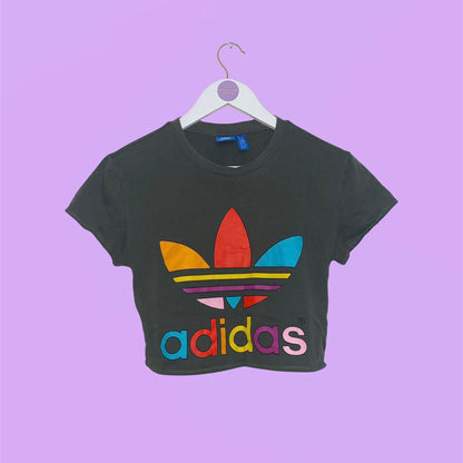 khaki crop top with short sleeves and big rainbow adidas text and symbol shown on a white clothes hanger on a lilac background