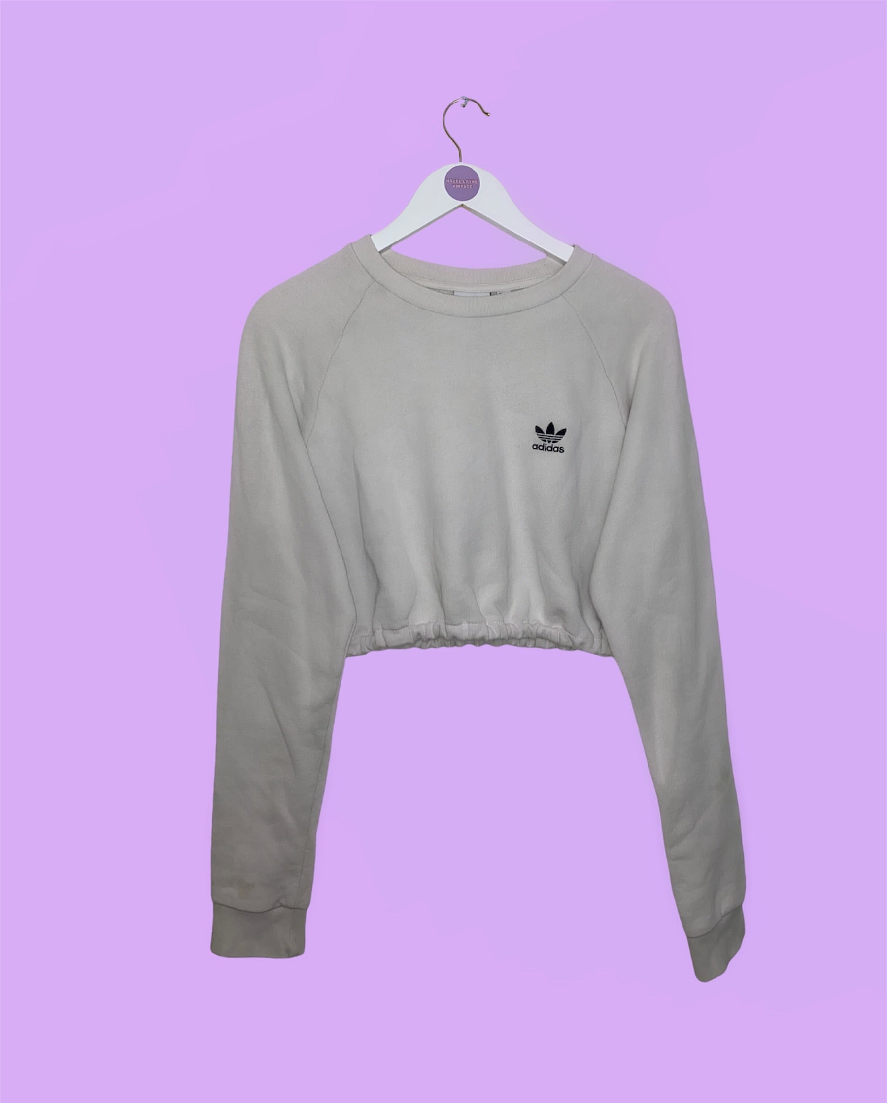 white cropped sweatshirt with black small adidas logo on chest shown on a white clothes hanger on a lilac background
