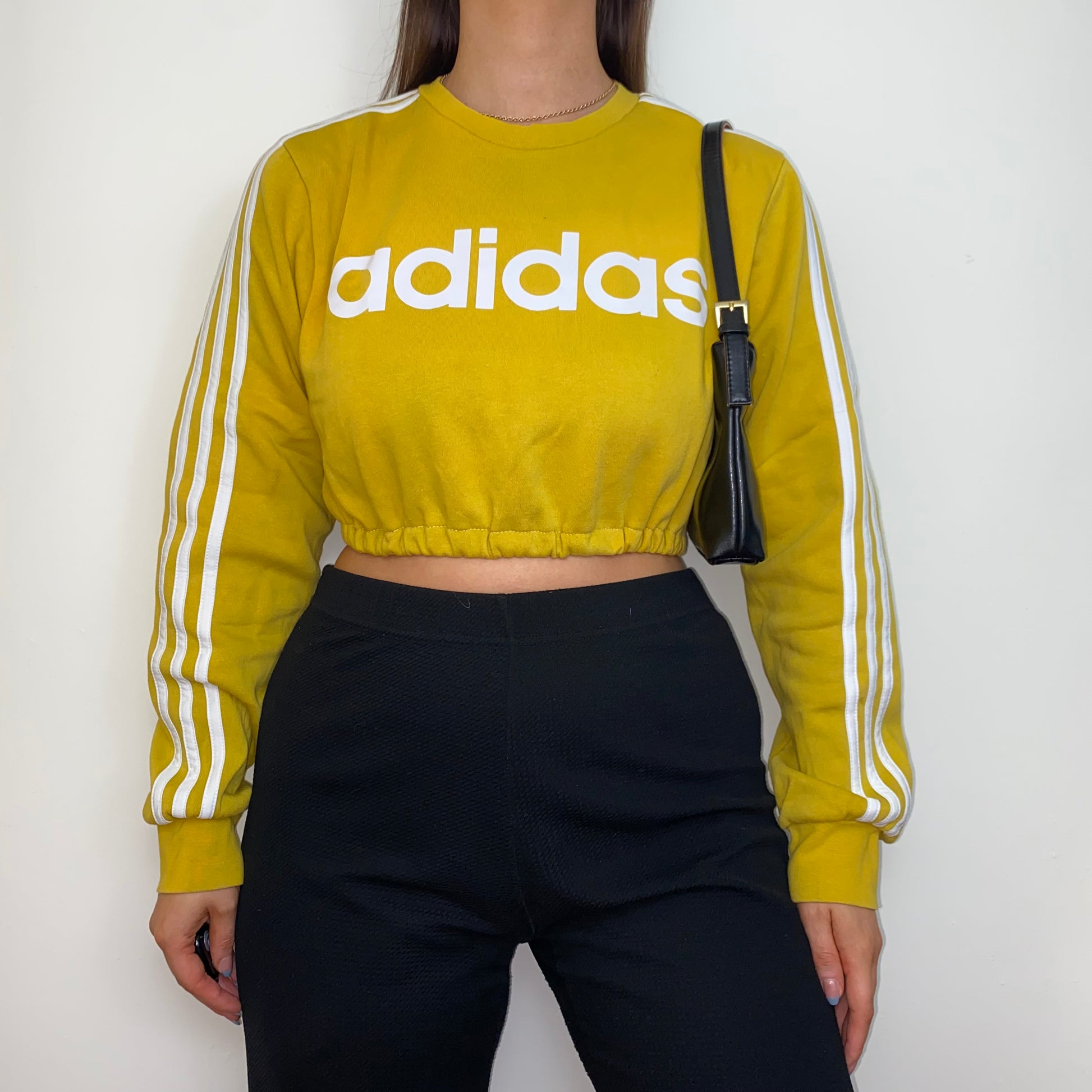 yellow cropped sweatshirt with white adidas logo shown on a model wearing black trousers and a black shoulder bag
