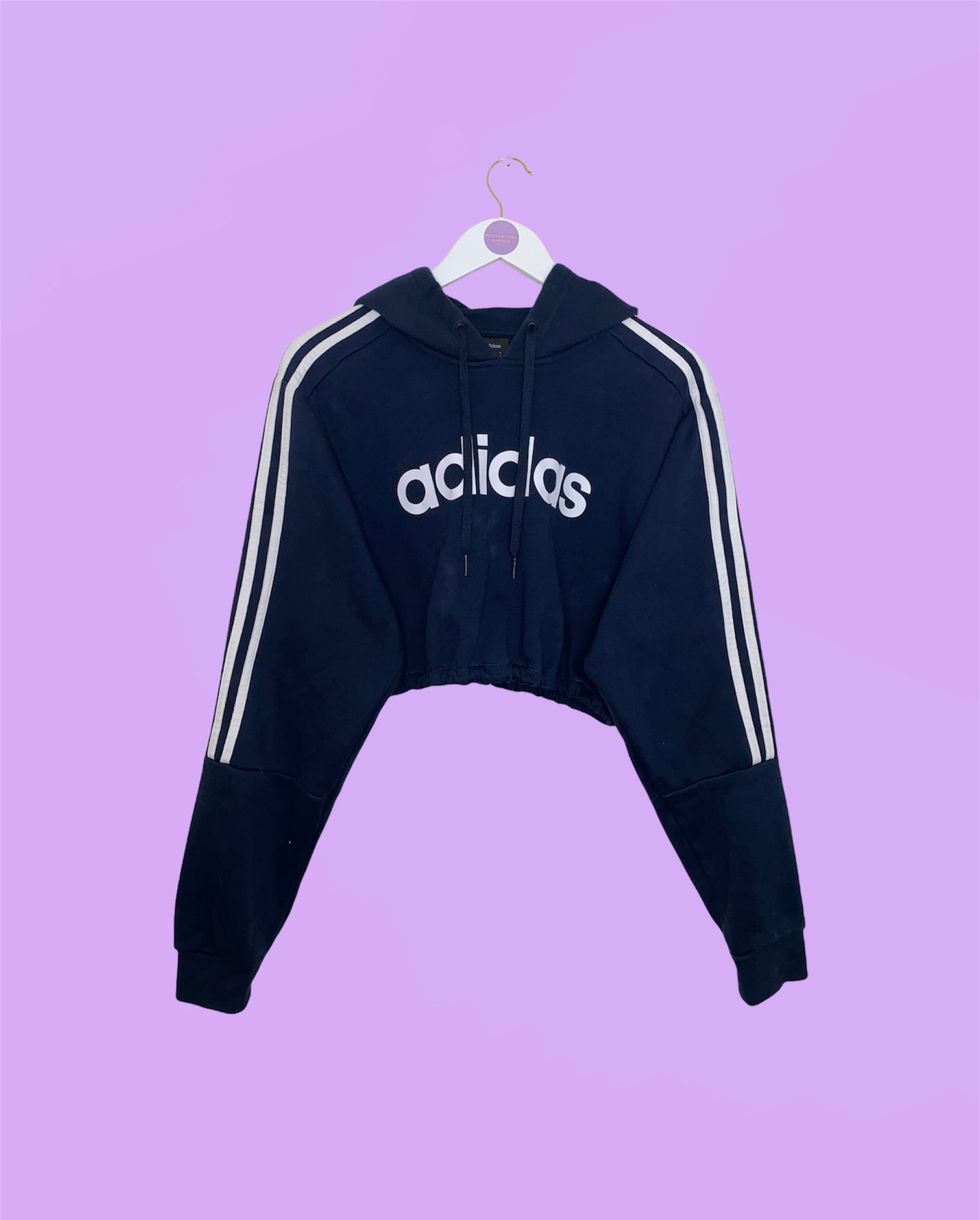 navy adidas cropped hoodie with white text adidas logo and stripes on sleeves shown on a white clothes hanger on a lilac background