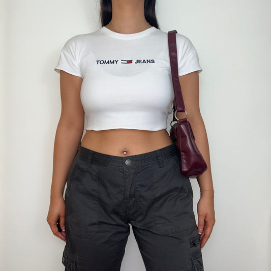 white short sleeve crop top with black tommy jeans logo shown on a model wearing cargo trousers and a burgundy shoulder bag