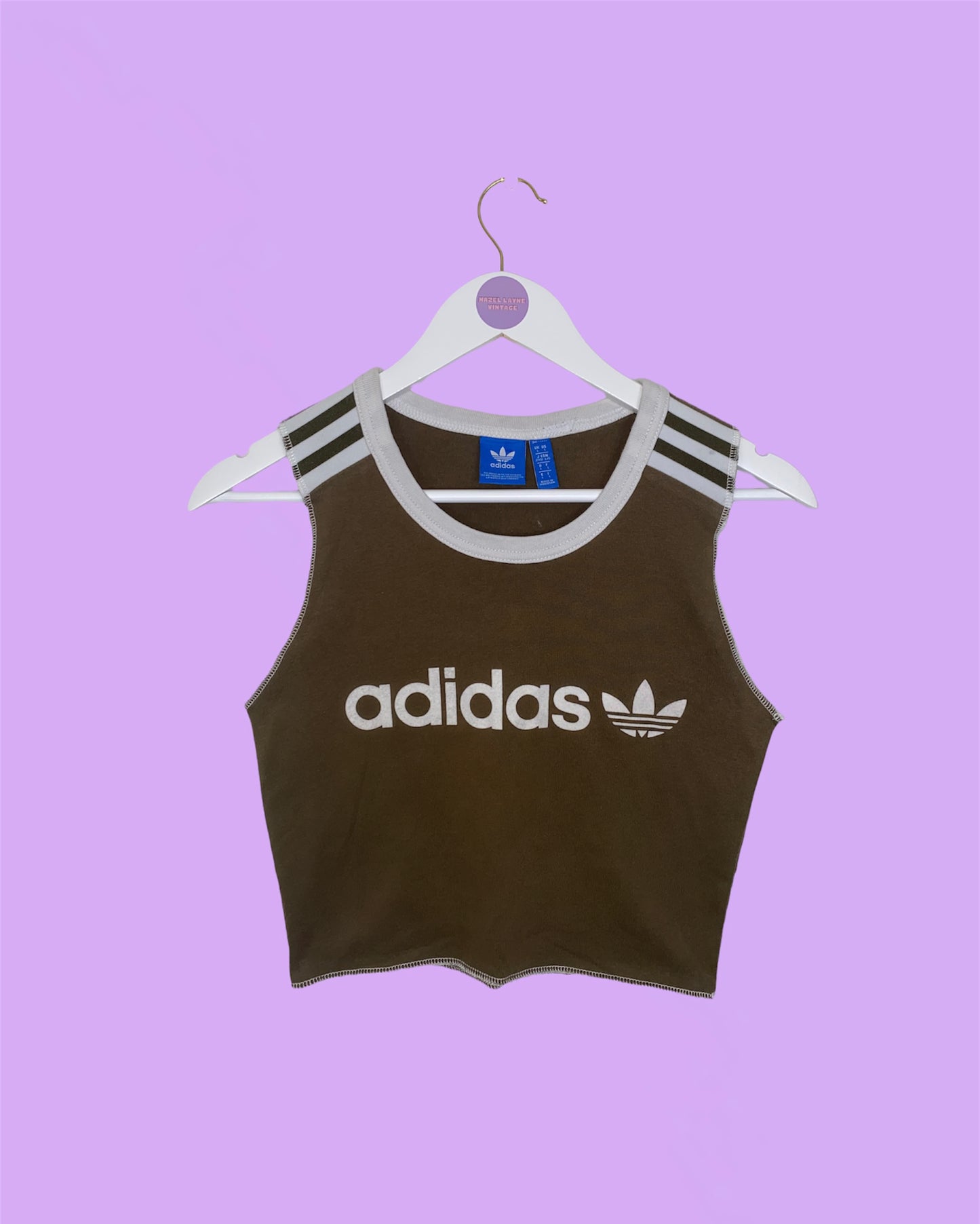 khaki sleeveless crop top with white adidas logo shown on a white clothes hanger on a lilac background