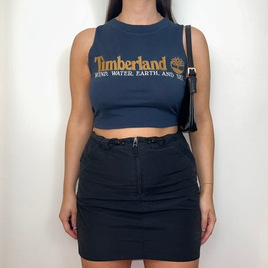 navy sleeveless crop top with orange and white timberland logo shown on a model wearing a black mini skirt and black shoulder bag