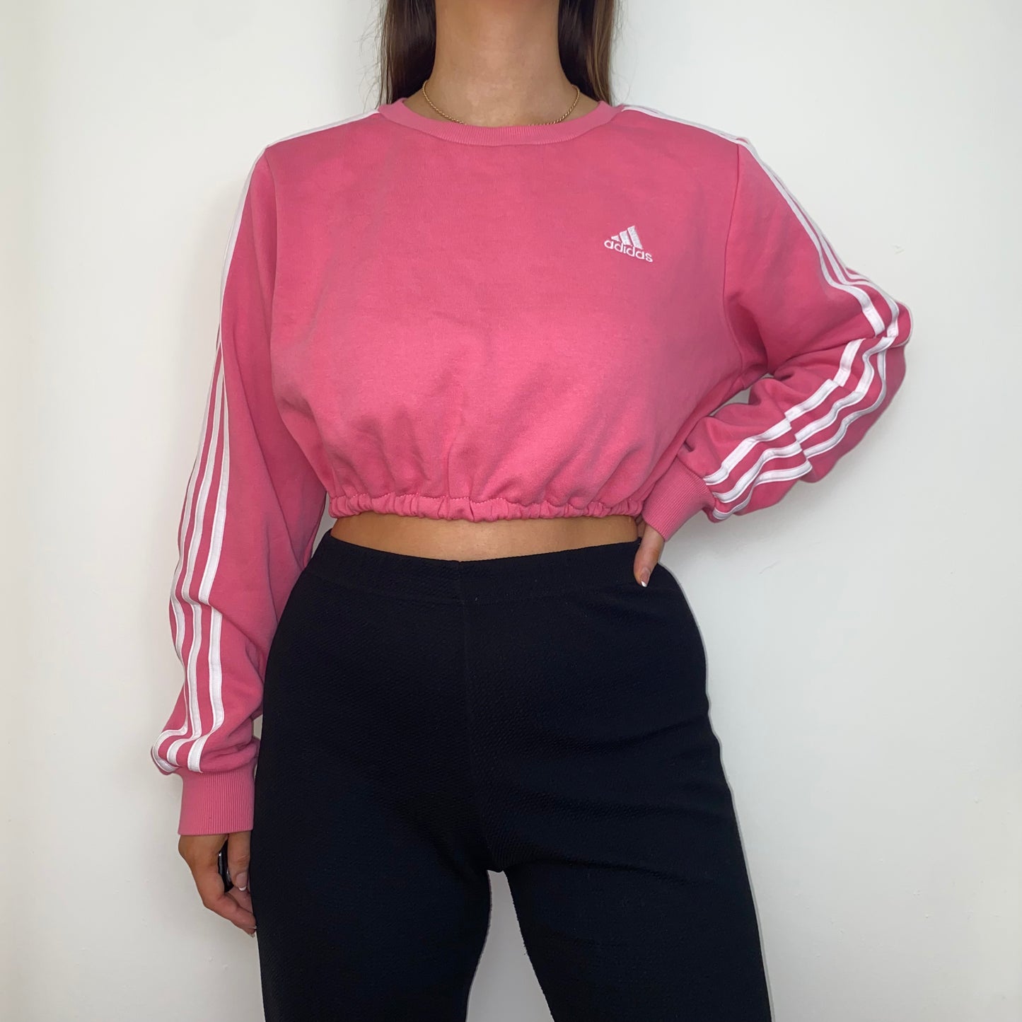 pink adidas cropped sweatshirt with white adidas logo shown on a model wearing black trousers with hand on hip