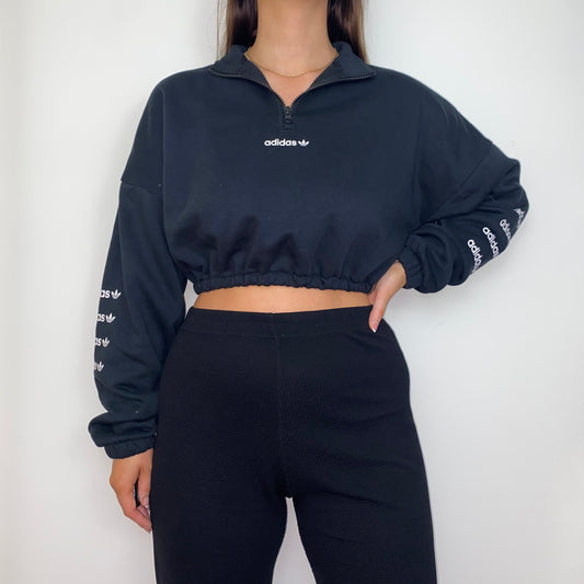 black 1/4 zip cropped sweatshirt with white adidas logo on front and sleeves shown on a model wearing black trousers