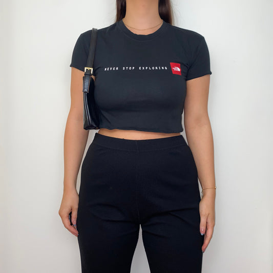 black short sleeve crop top with white and red north face logos shown on a model wearing black trousers and a black shoulder bag
