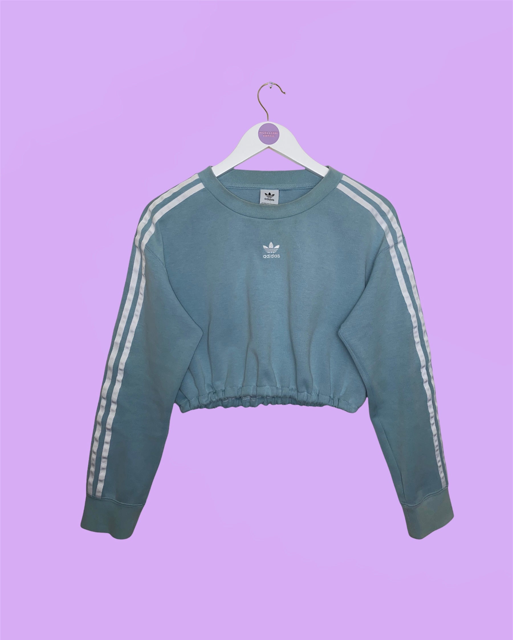 light blue cropped sweatshirt with white adidas logo on chest and 3 stripes on sleeves shown on a white clothes hanger on a lilac background