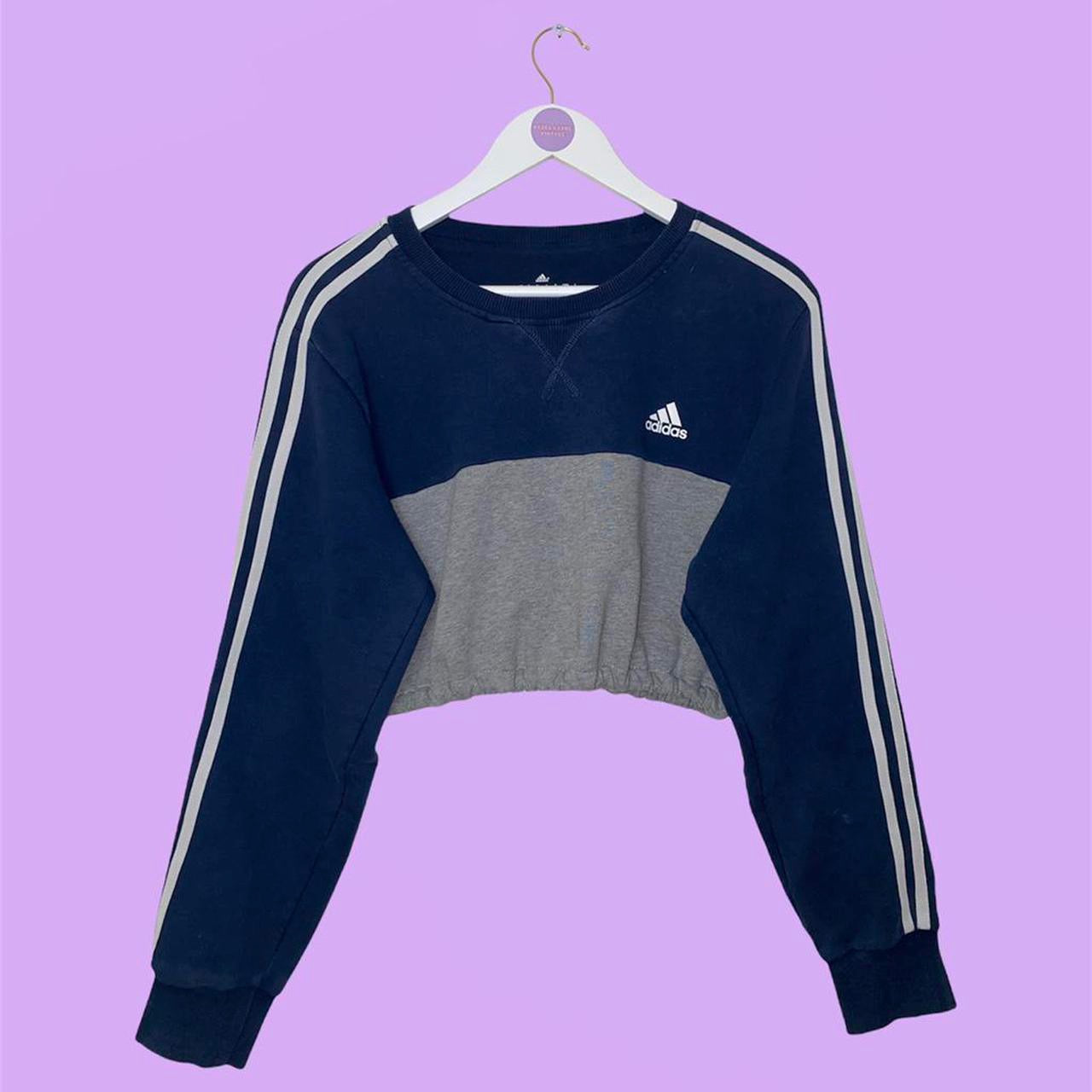 navy and grey cropped sweatshirt with white adidas text and symbol logo on chest with 3 stripes on sleeves shown on a white clothes hanger on a lilac background