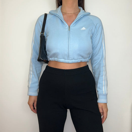 light blue 1/4 zip cropped sweatshirt with white adidas logo shown on a model wearing black trousers and a black shoulder bag