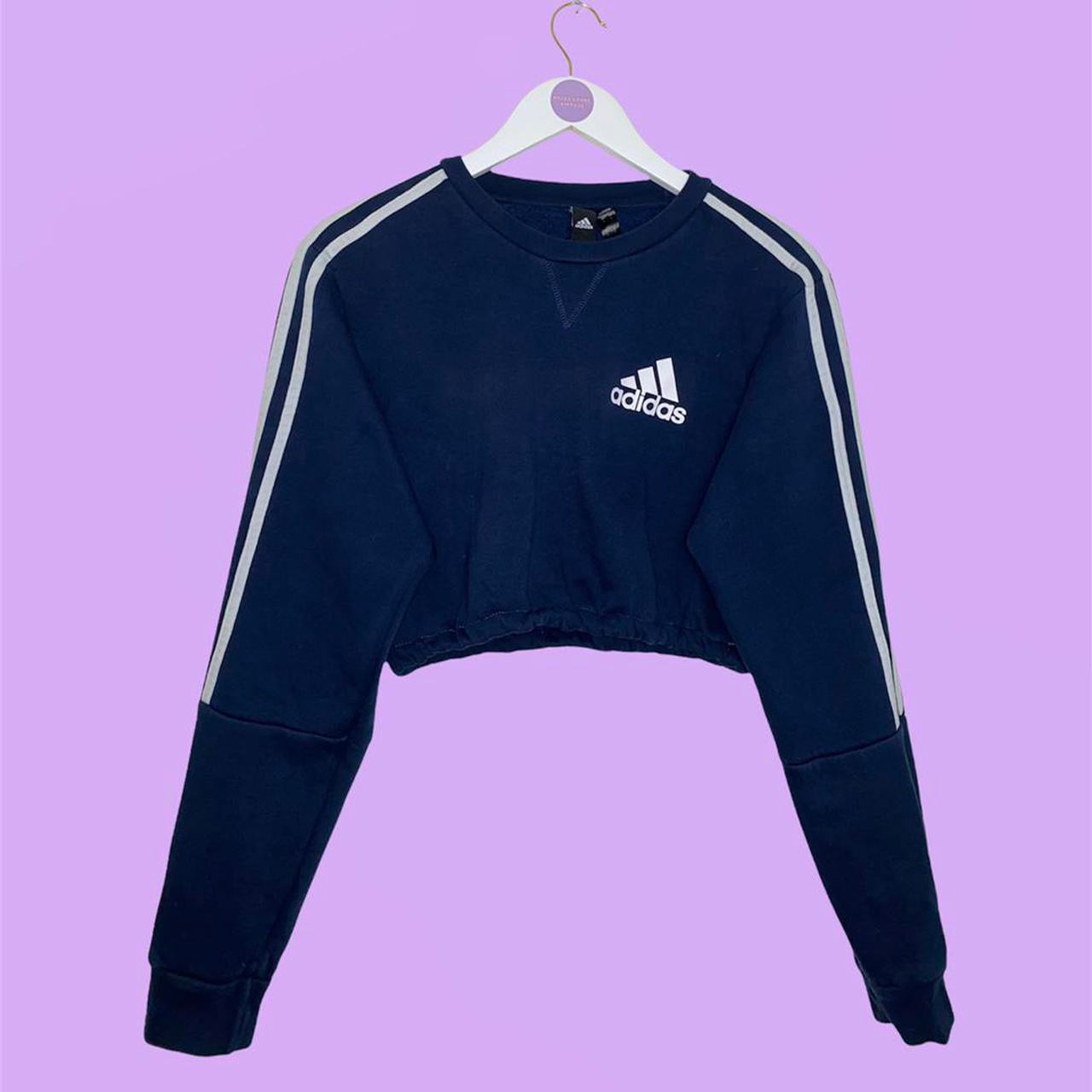 navy sweatshirt with small adidas text and symbol logo on chest with white 3 stripes on arms shown on white clothes hanger on a lilac background