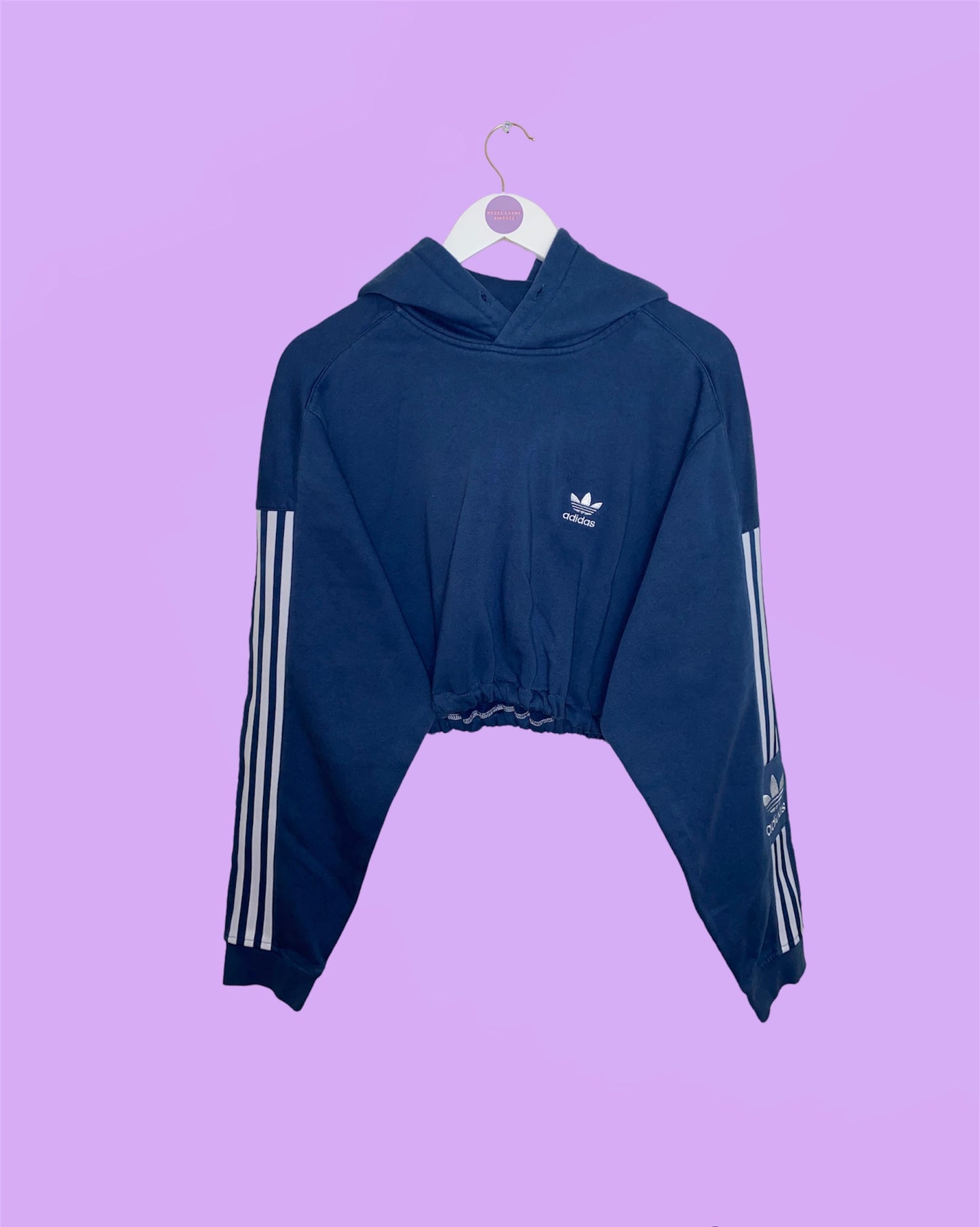 blue cropped hoodie with white adidas logo on chest and sleeves shown on a white clothes hanger on a lilac background