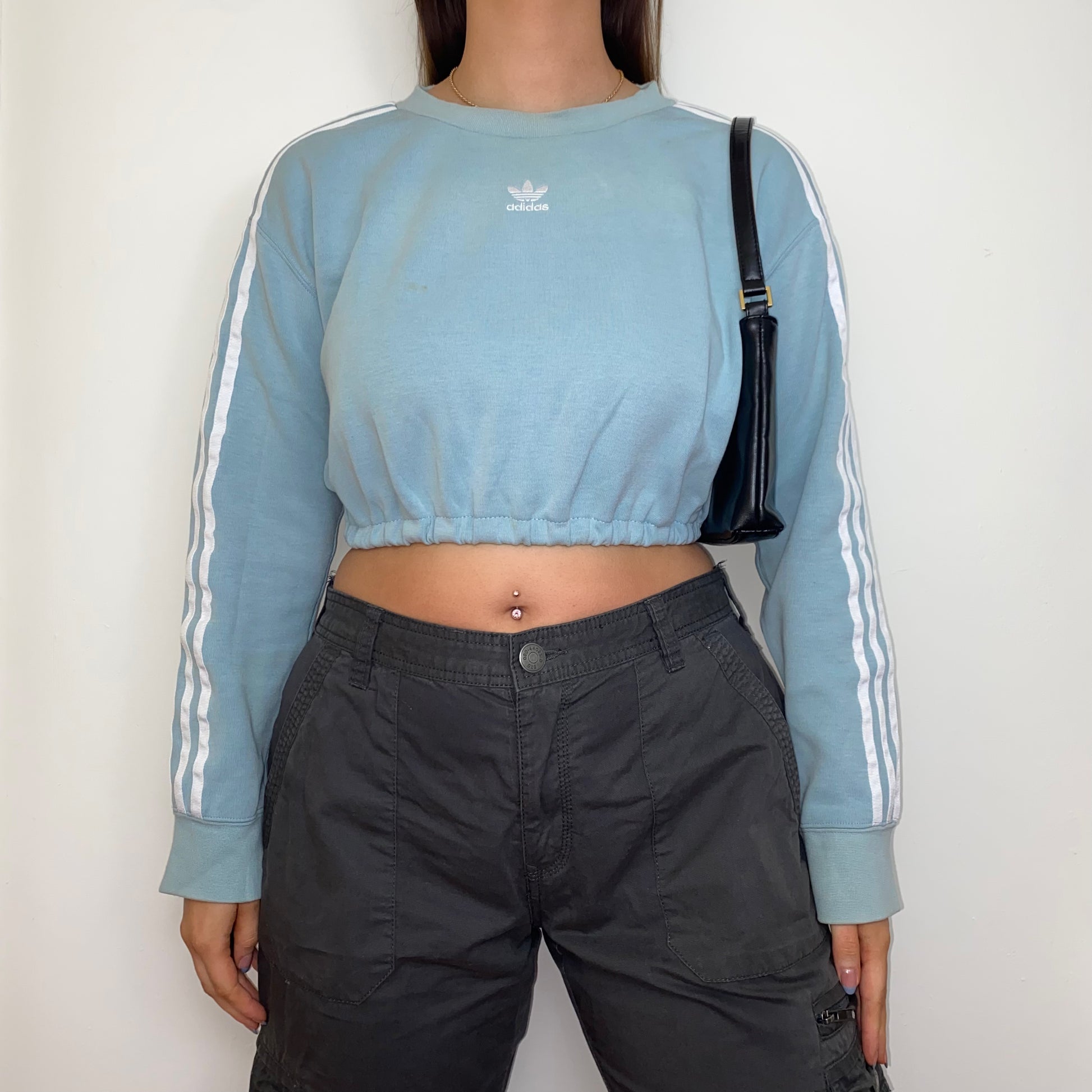 light blue cropped sweatshirt with white adidas logo shown on a model wearing grey cargo trousers and a black shoulder bag