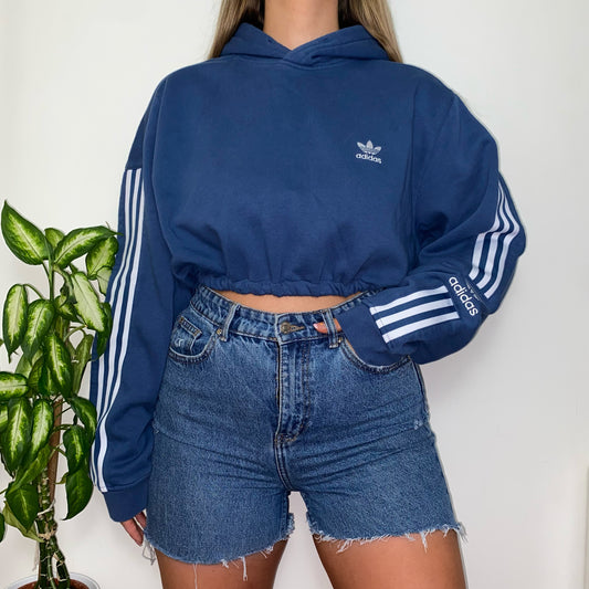 blue cropped hoodie with white adidas logo on chest and sleeves shown on a model wearing blue denim shorts with hand in pocket